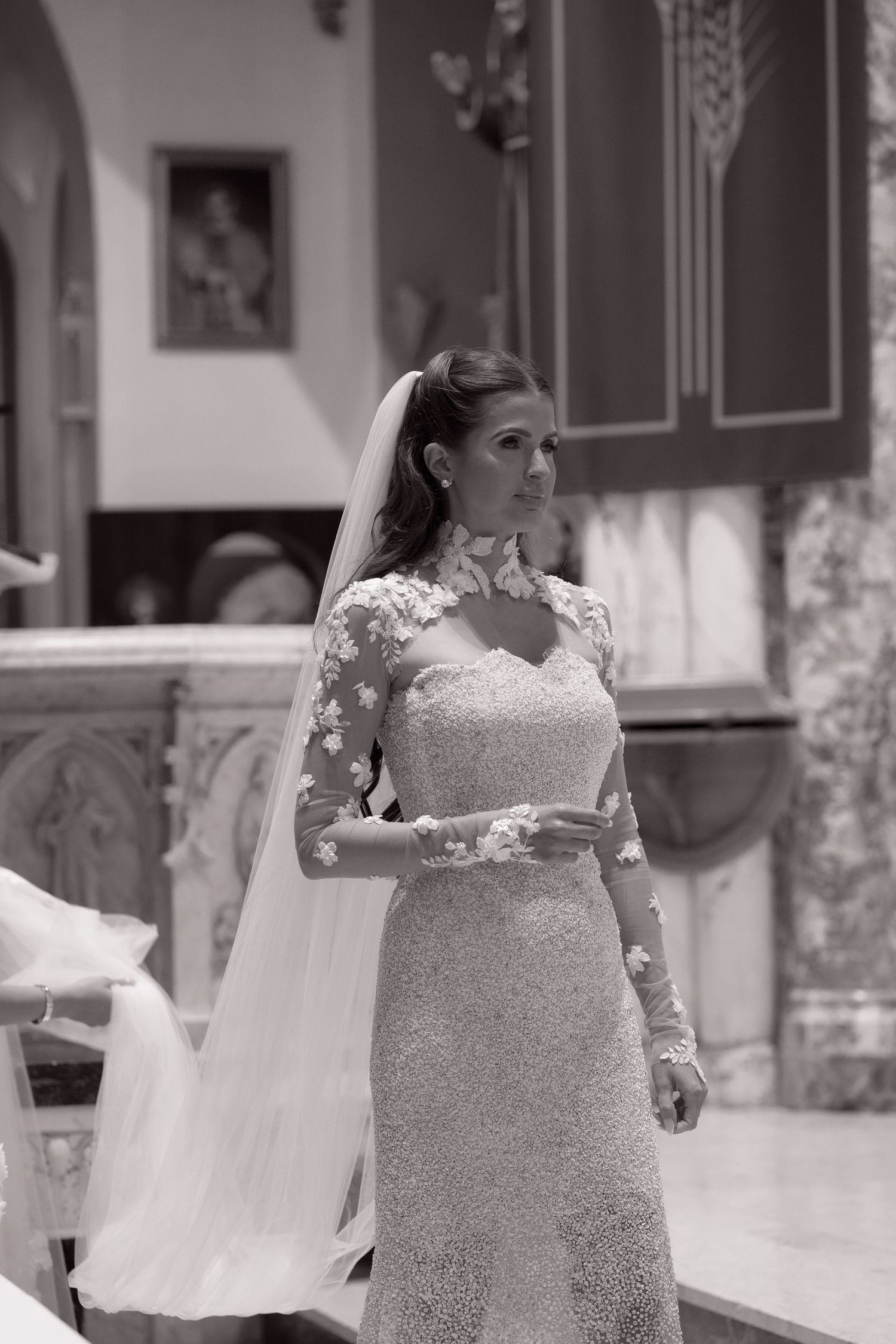 Elegant bride in a long-sleeve lace wedding gown with floral appliques and veil stands at the church altar.