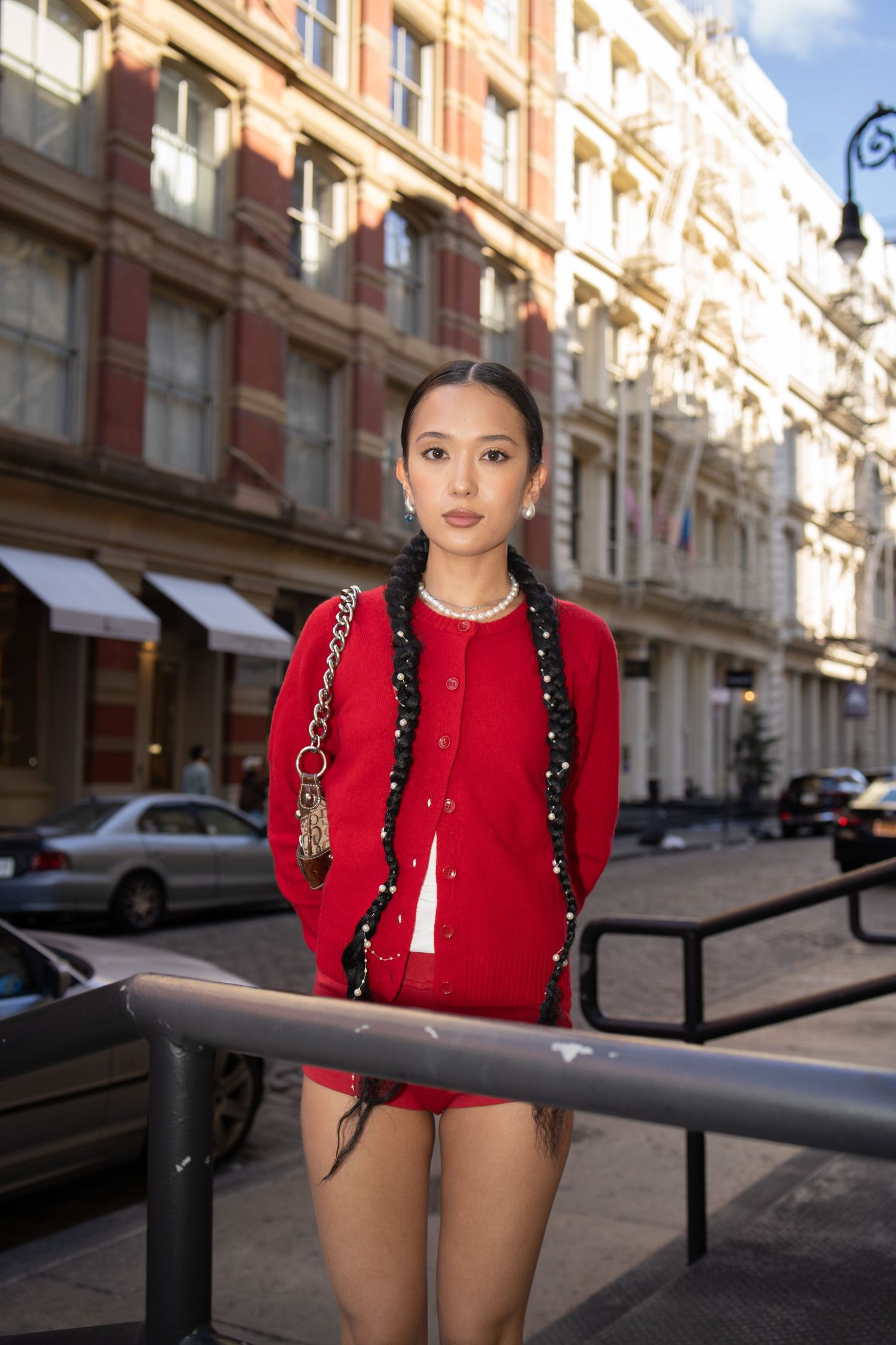 Stylish woman in red cardigan and skirt with braided hair stands on urban street near railing.