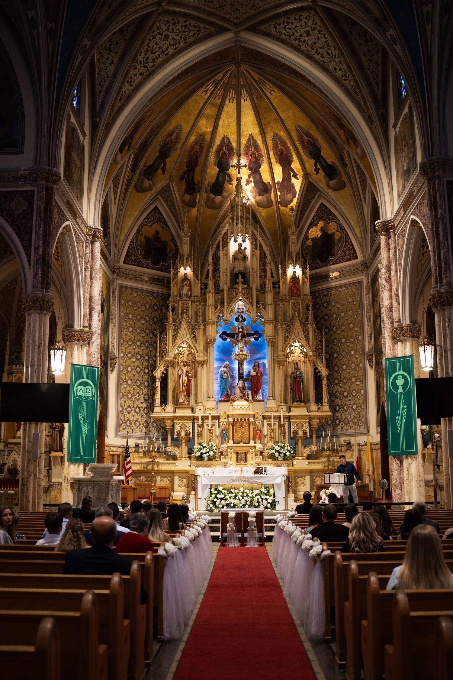 Grand ornate Catholic church interior with red carpet aisle, decorated pews, and golden altar during wedding.