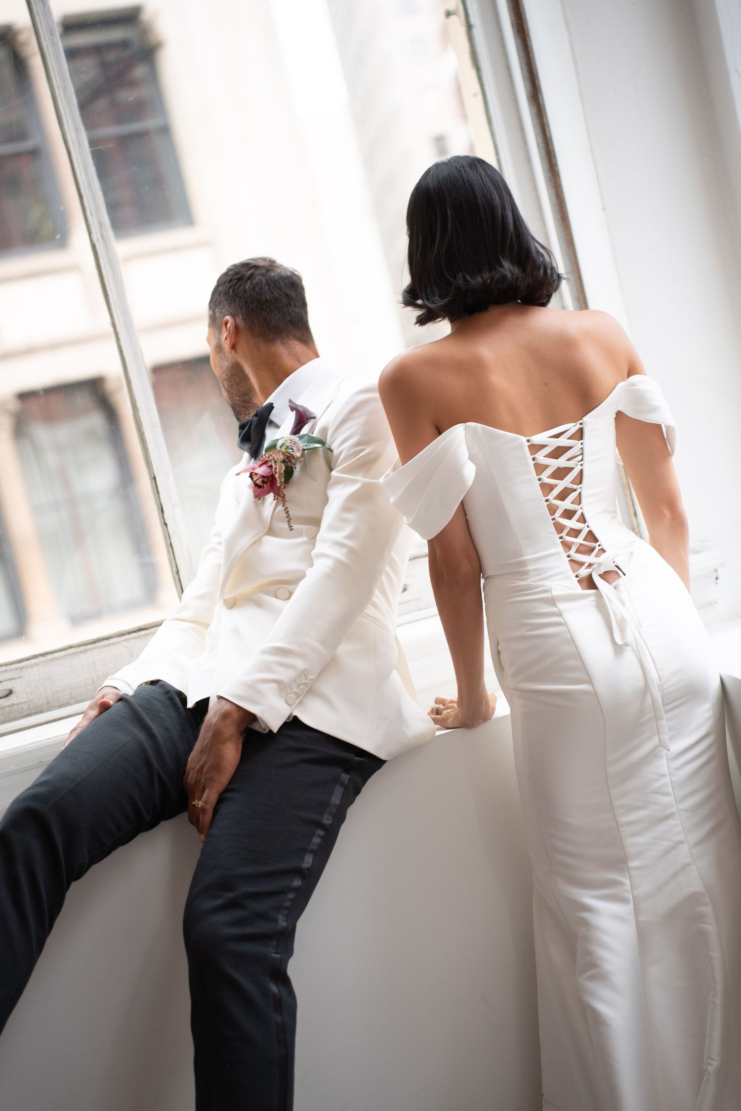 Bride and groom standing at window, bride wearing elegant off-shoulder lace-back wedding gown.