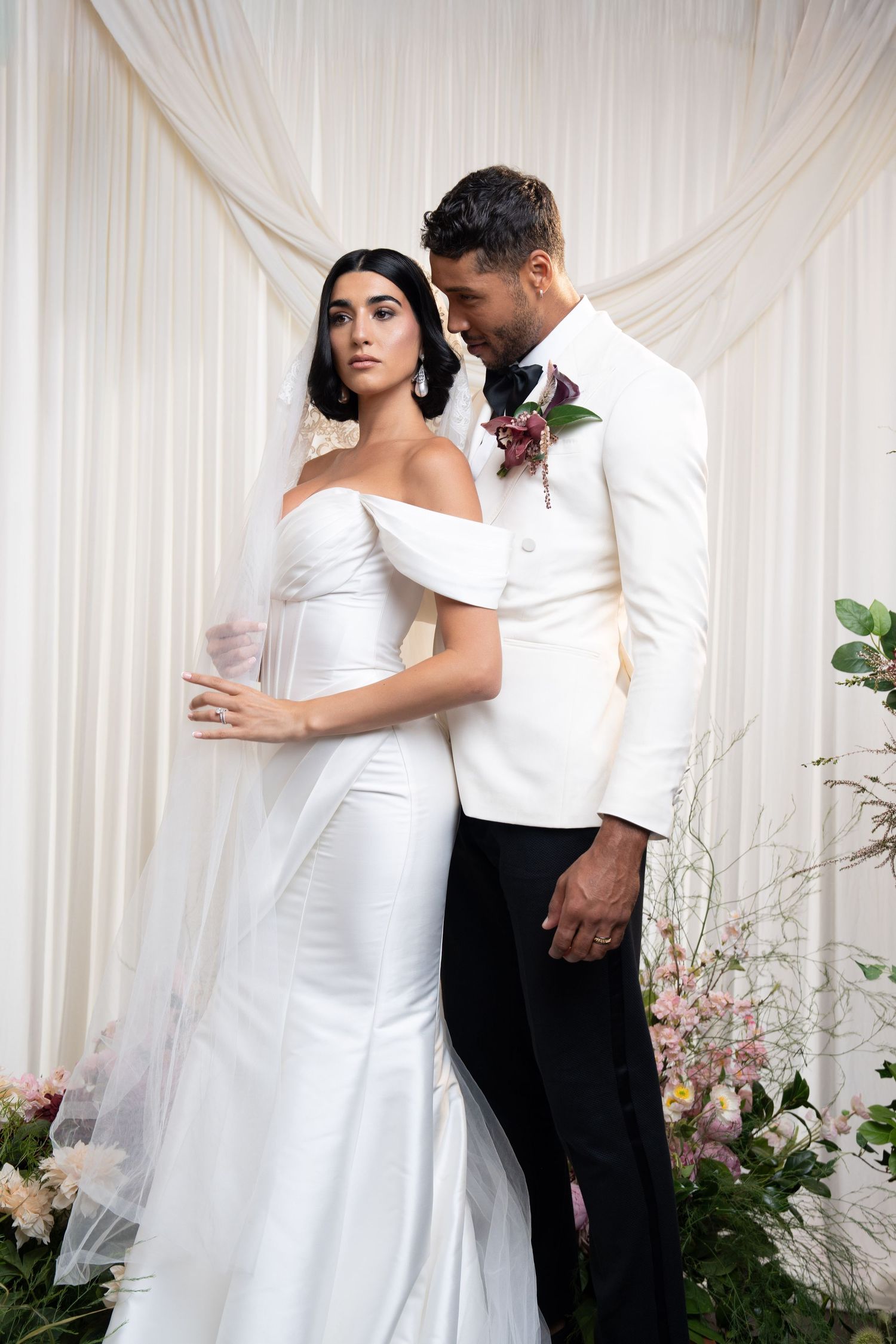 Groom in white tuxedo embraces bride in off-shoulder gown near floral arrangements and white drapes.