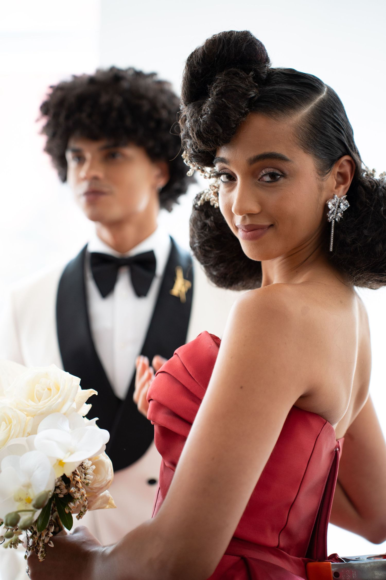 Smiling woman in red strapless dress with sculptural updo and chandelier earrings poses with white floral bouquet.