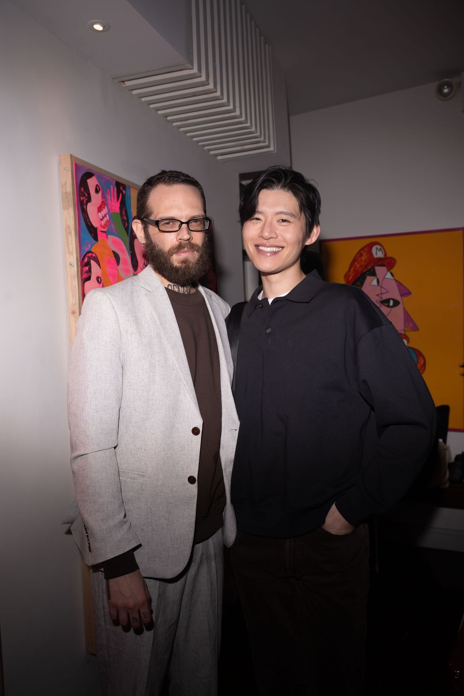Two men pose together smiling beside vibrant pop art paintings at gallery.