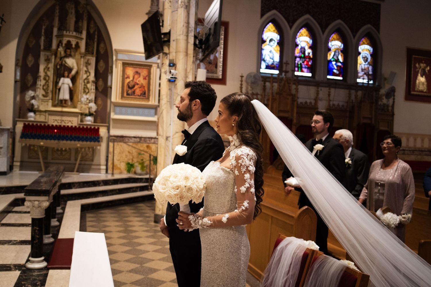 Bride in lace gown with long veil and groom in tuxedo stand together during church wedding ceremony.