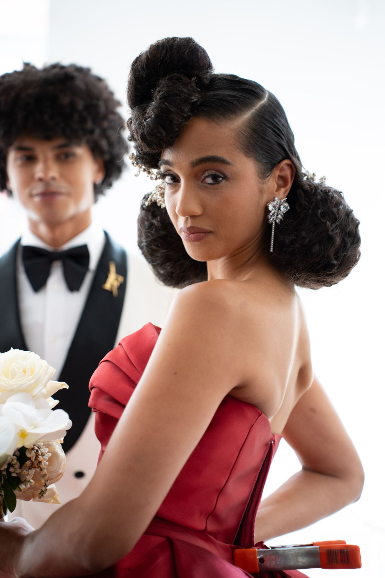 Elegant bride in red strapless gown with sculptural updo and diamond earrings holding white roses at wedding.