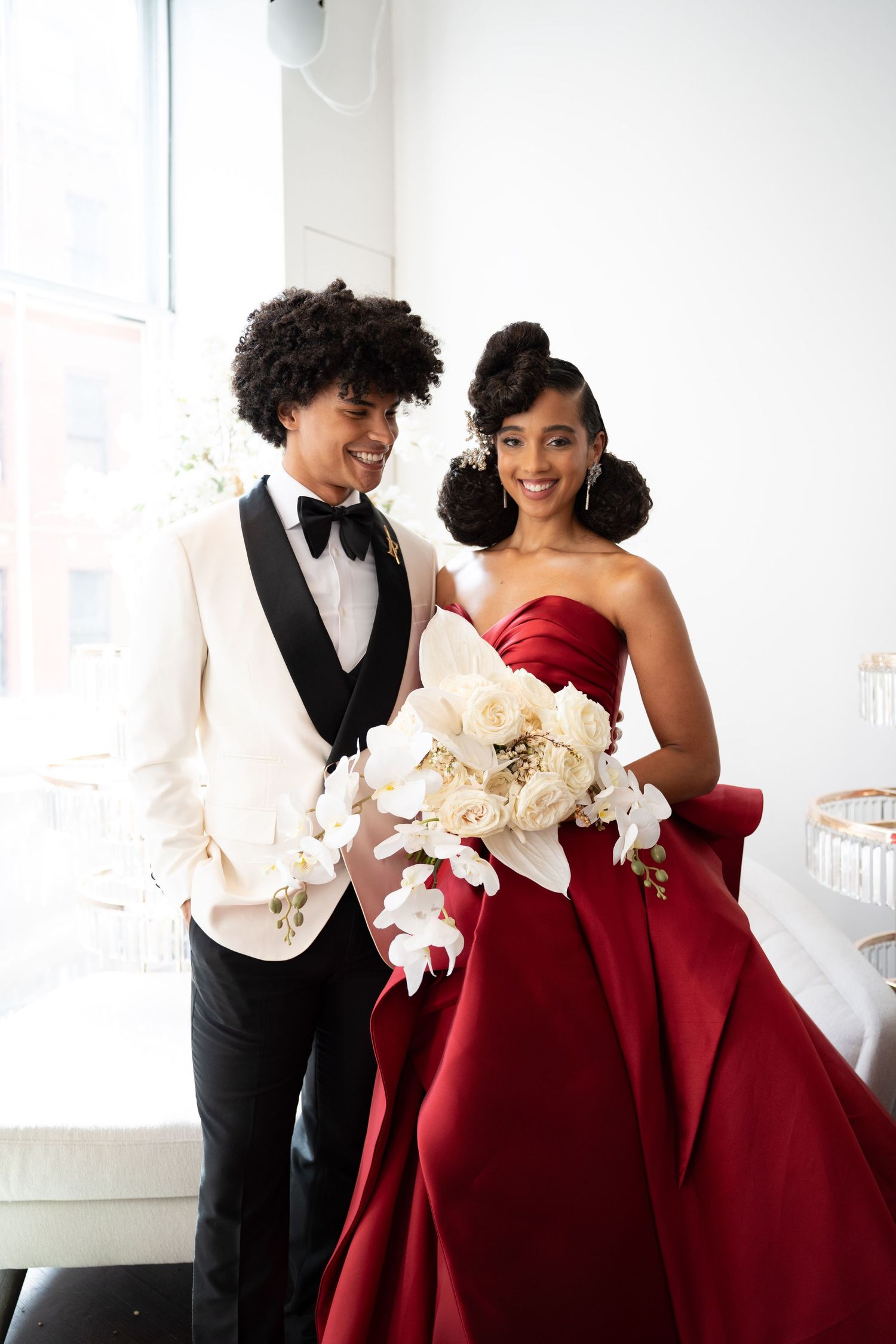 Happy wedding couple posing, groom in white tuxedo, bride in flowing red satin gown holding elegant white floral bouquet.