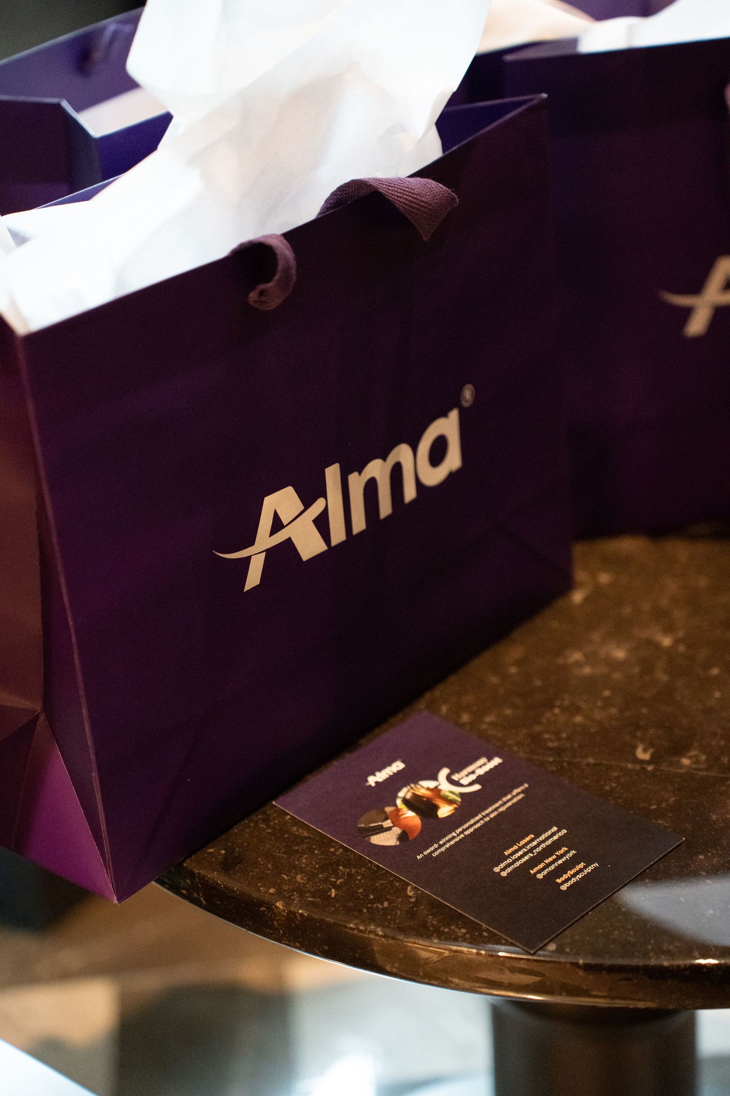 Purple Alma branded gift bags with tissue paper on a marble table at a beauty event.