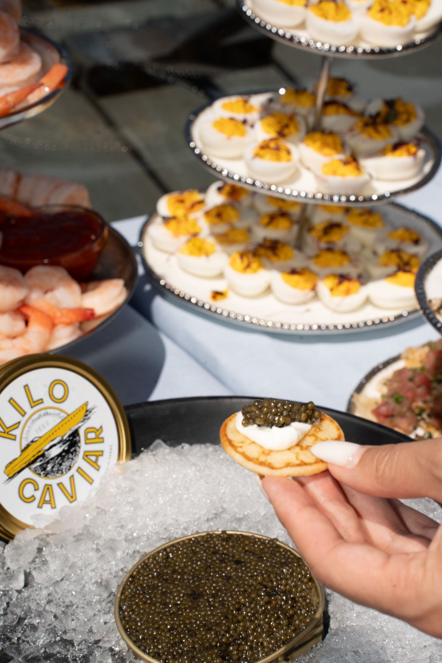 Hand holding blini with Kilo Caviar beside deviled eggs tower and shrimp at luxury outdoor tasting event.