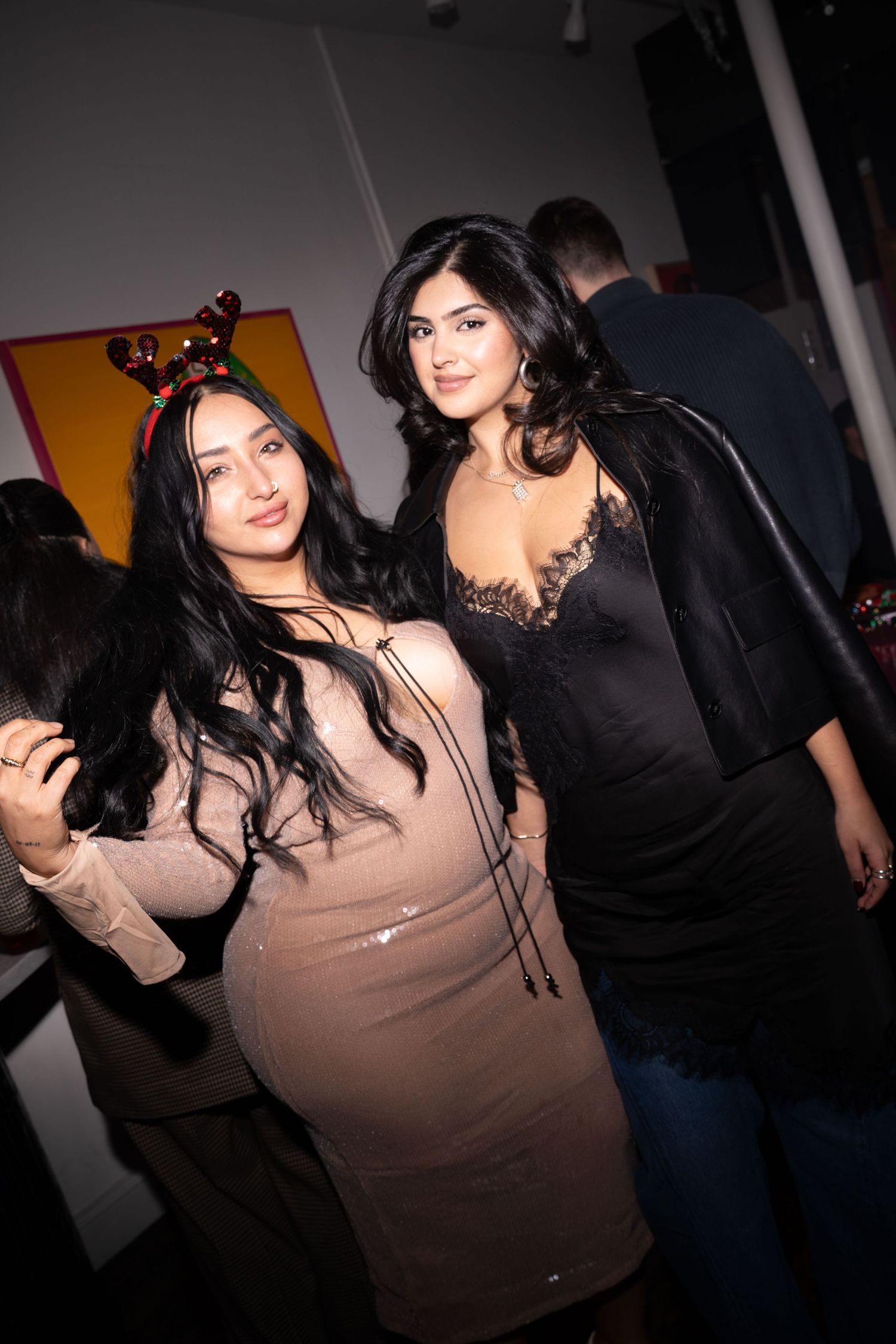 Two women in elegant party dresses pose together at an indoor social event.