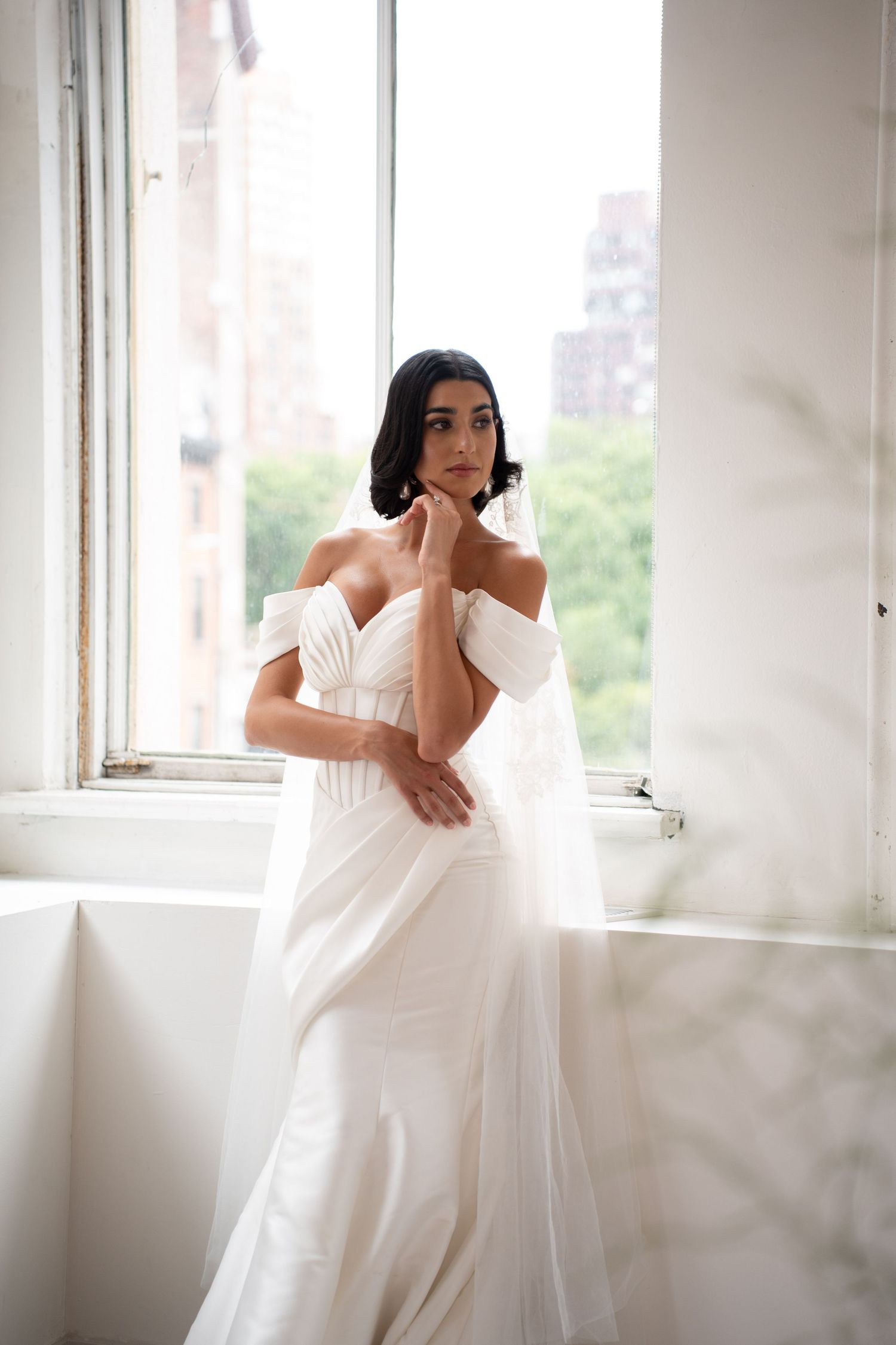 Thoughtful bride in off-shoulder white wedding gown with veil gazing out sunlit window.