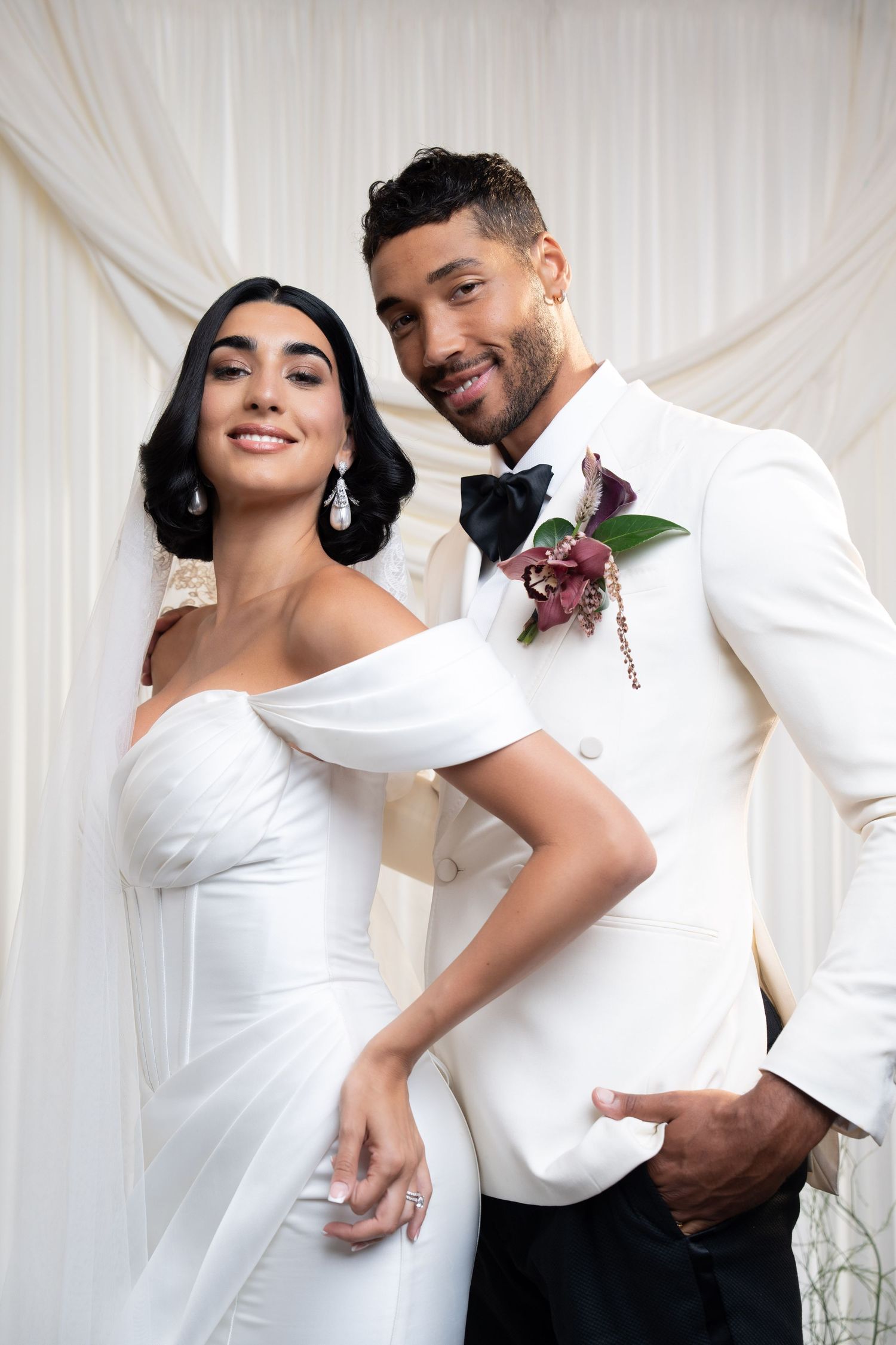 Smiling bride and groom in white wedding attire pose together against draped white fabric backdrop.