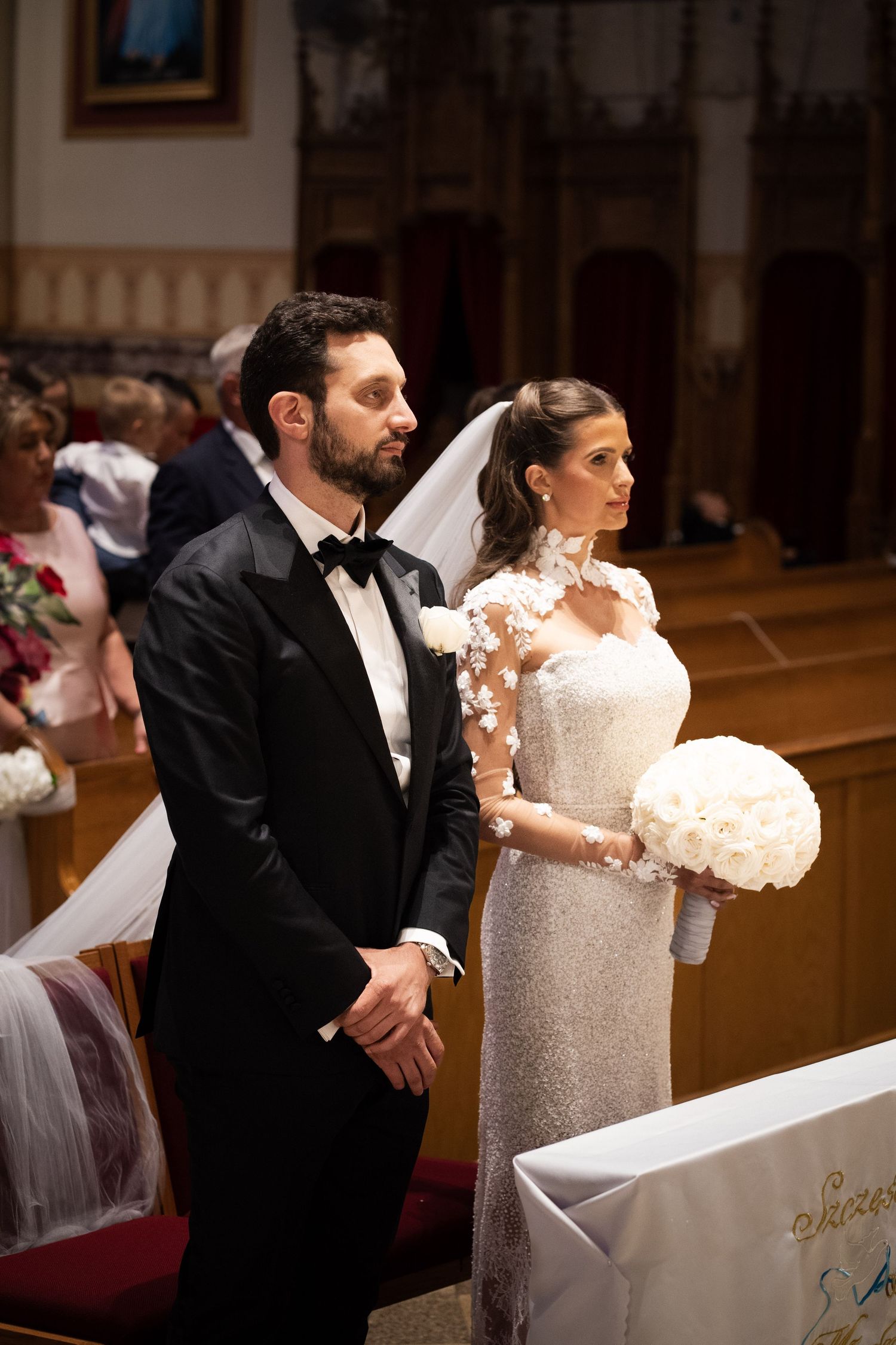 Bride in lace long-sleeve gown holding white rose bouquet stands with tuxedo-clad groom at church altar.