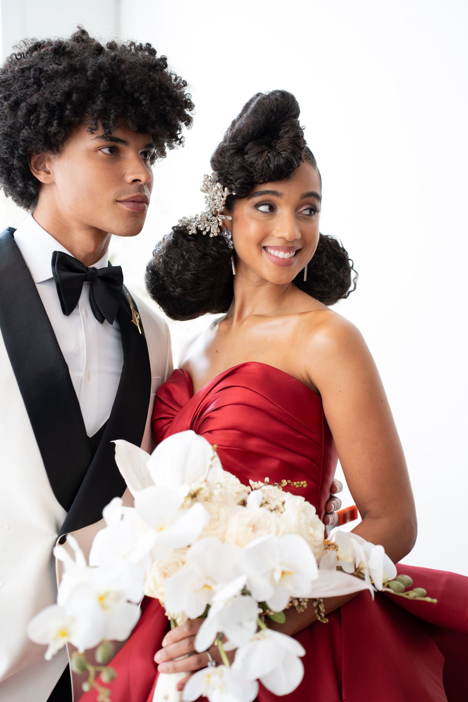 Smiling Black bride in red strapless gown with updo hairstyle, groom in white tuxedo holding white orchid bouquet.