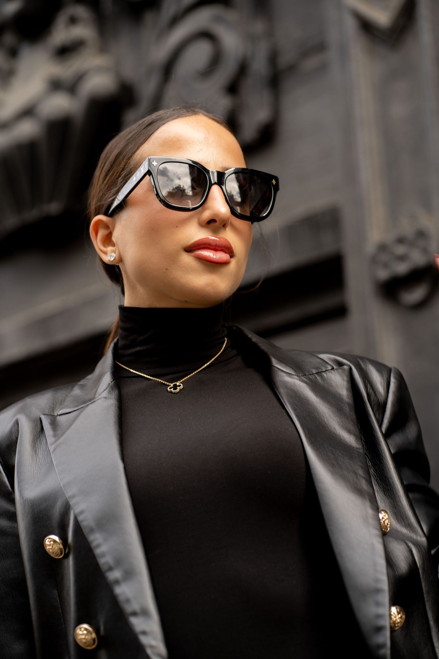 Close-up of woman wearing oversized black sunglasses, gold necklace, and black leather blazer with gold buttons.