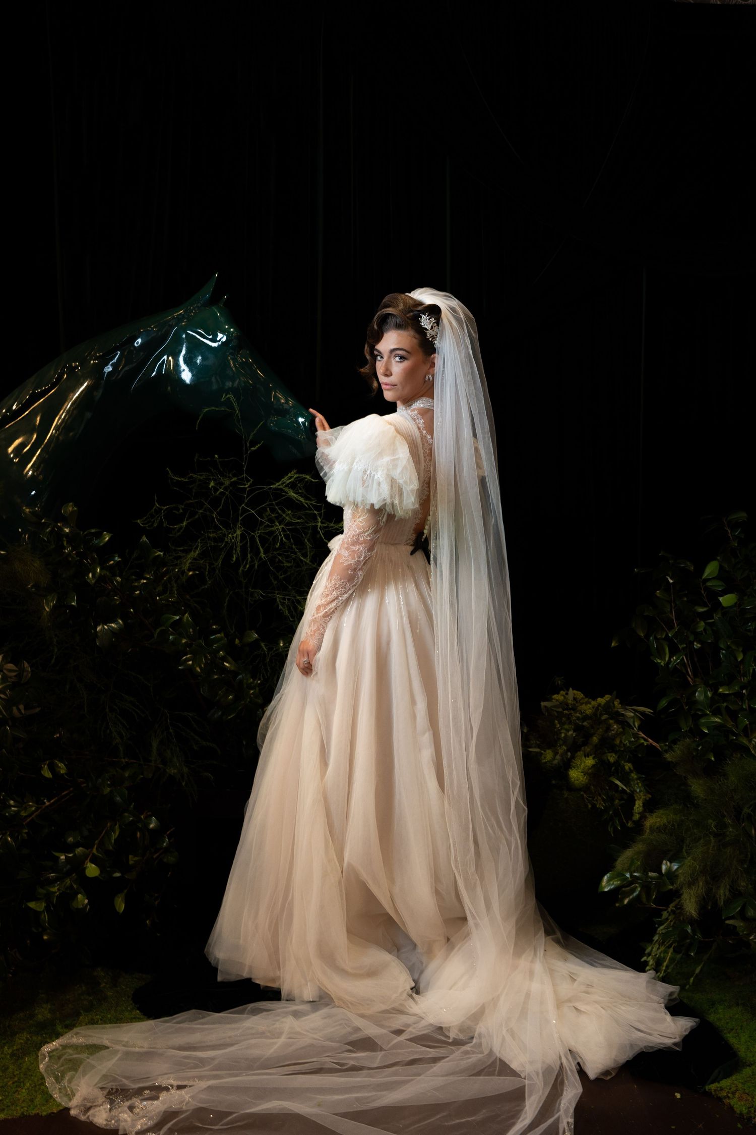 Bride in champagne tulle gown with veil poses beside dark horse sculpture among lush greenery.