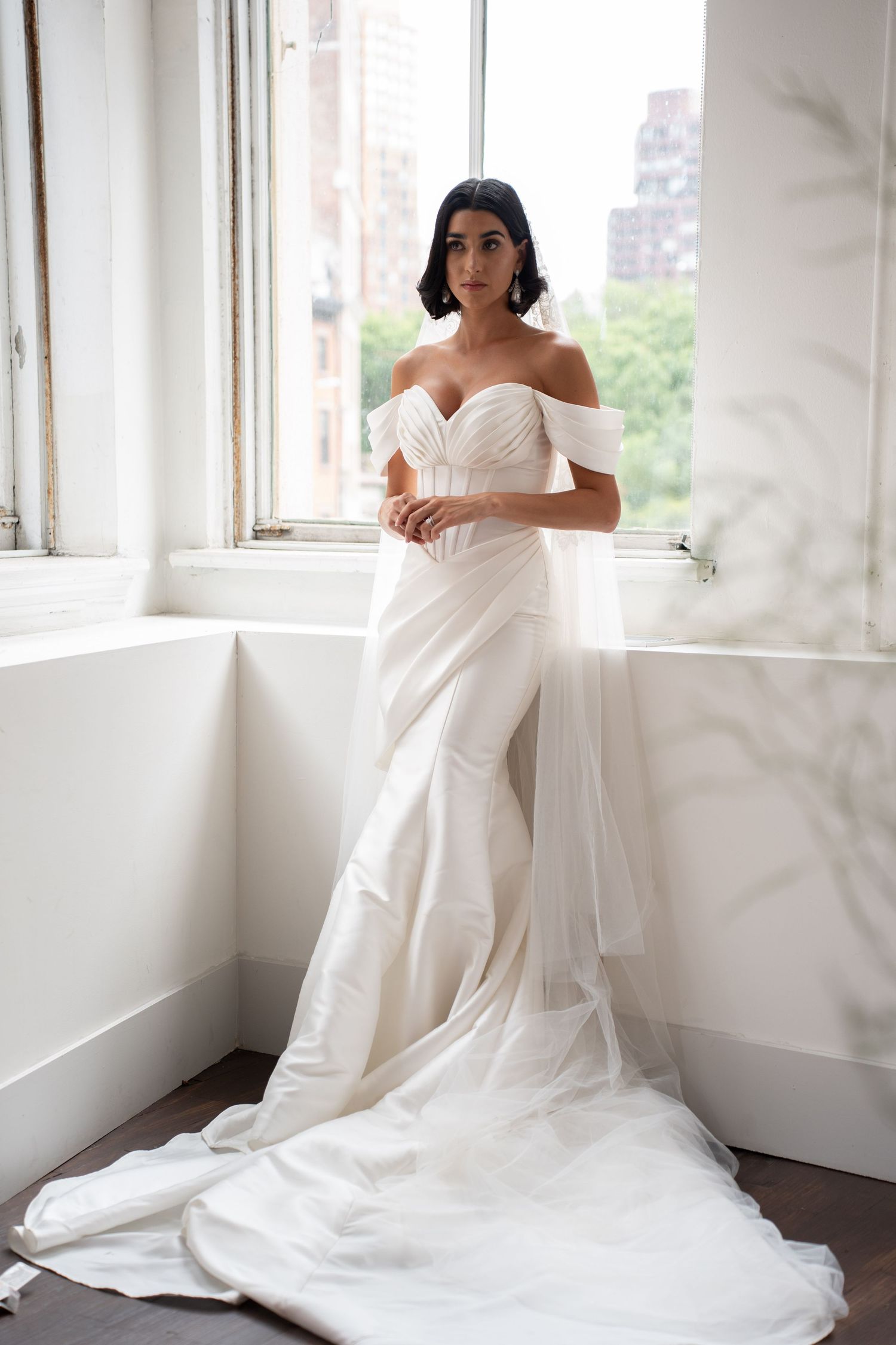 Bride in elegant off-shoulder white wedding gown with flowing veil standing by sunlit window.