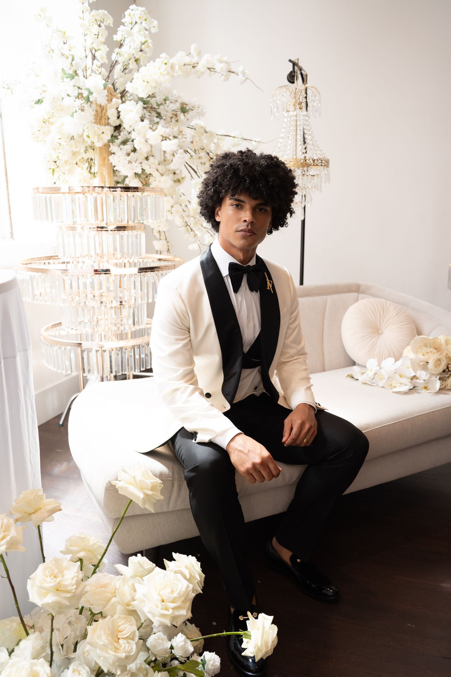 Groom in white tuxedo with black bow tie sits on cream sofa amid white roses at luxurious wedding venue.