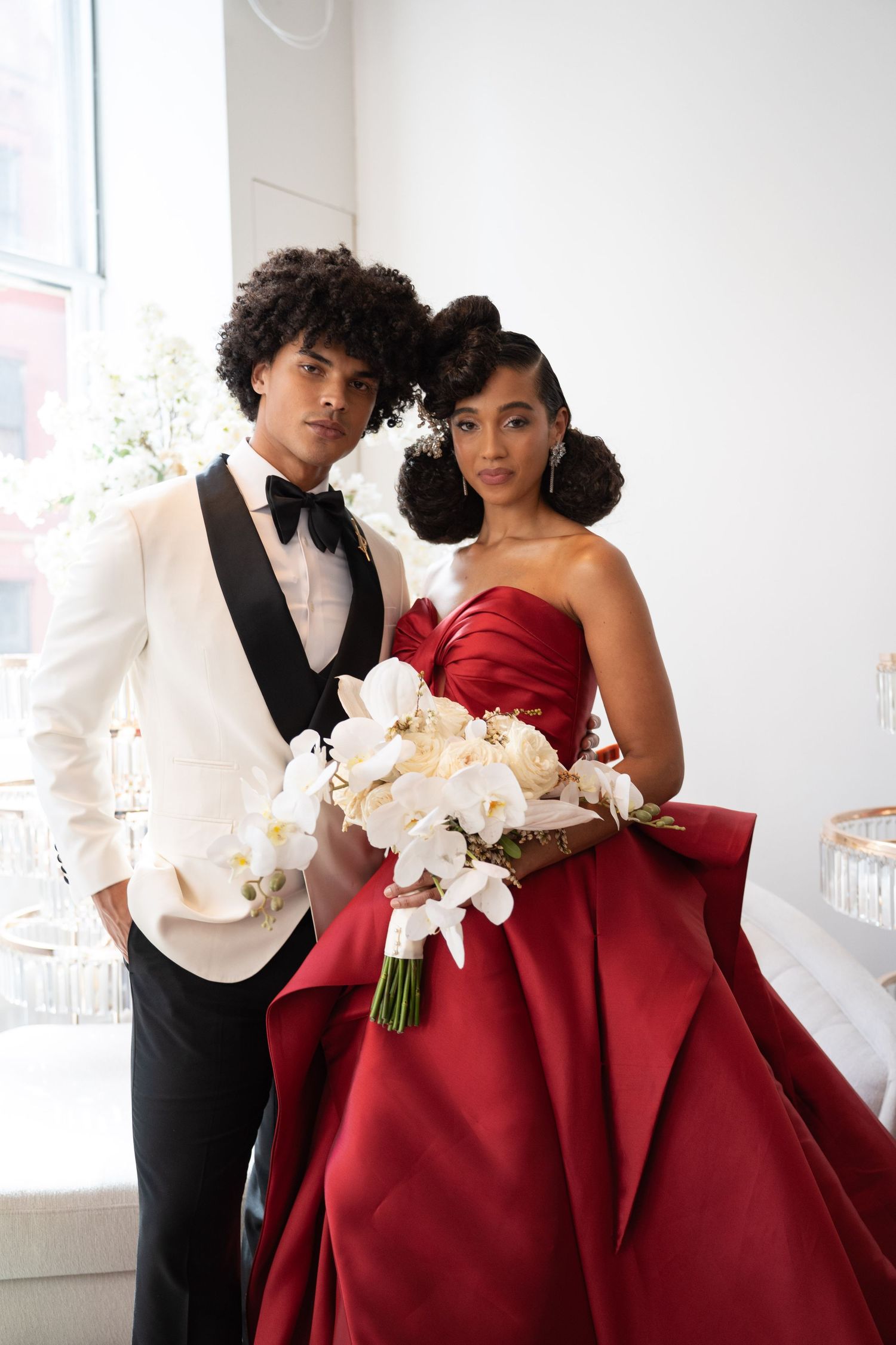 Black bride and groom posing formally, she holds white orchid bouquet wearing red strapless gown, he wears white tuxedo.
