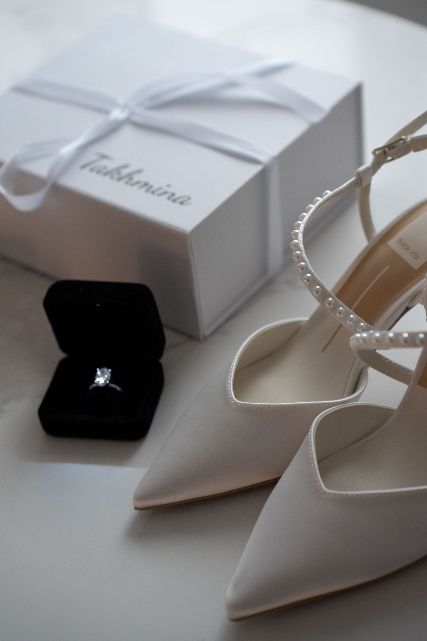 White bridal heels with pearl straps beside diamond ring in box and gift package.