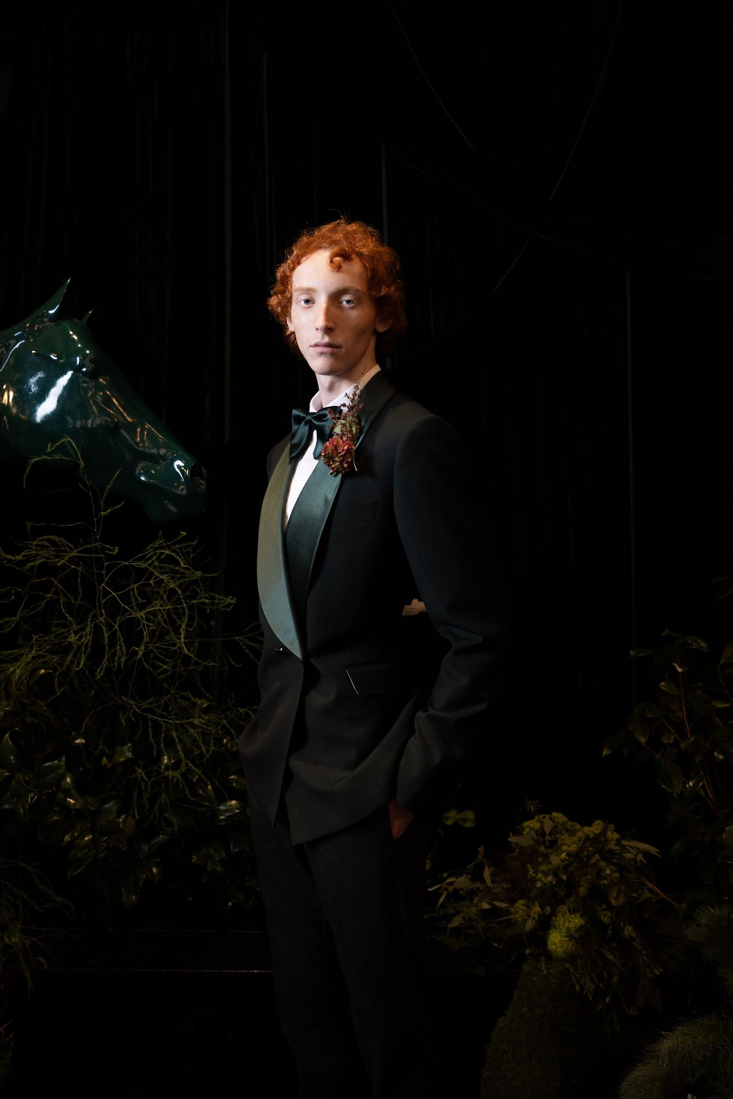 Red-haired groom in black tuxedo with floral boutonniere poses confidently among dark lush greenery.