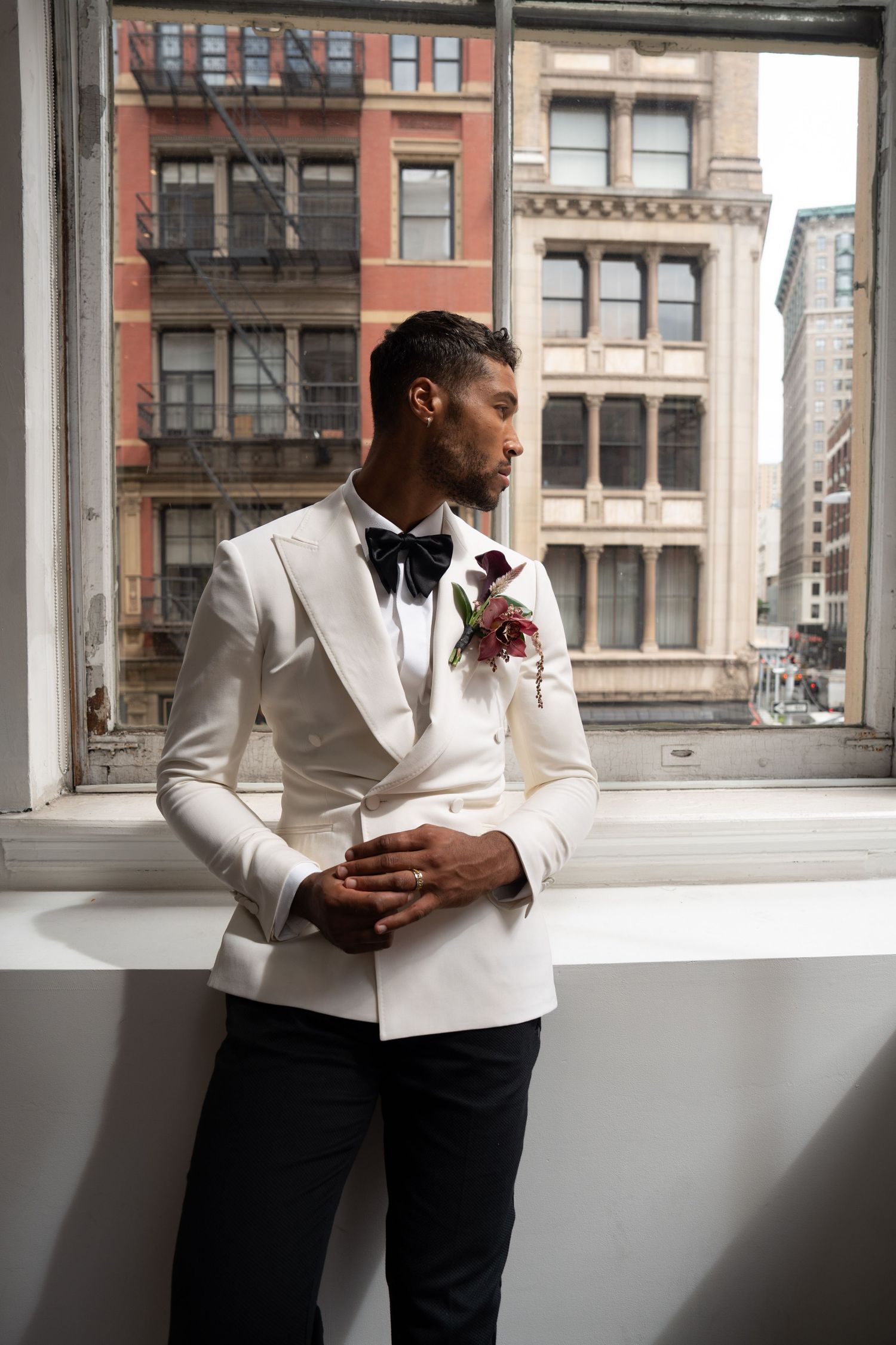 Groom in white tuxedo with black bow tie and burgundy boutonniere gazes out city window thoughtfully.