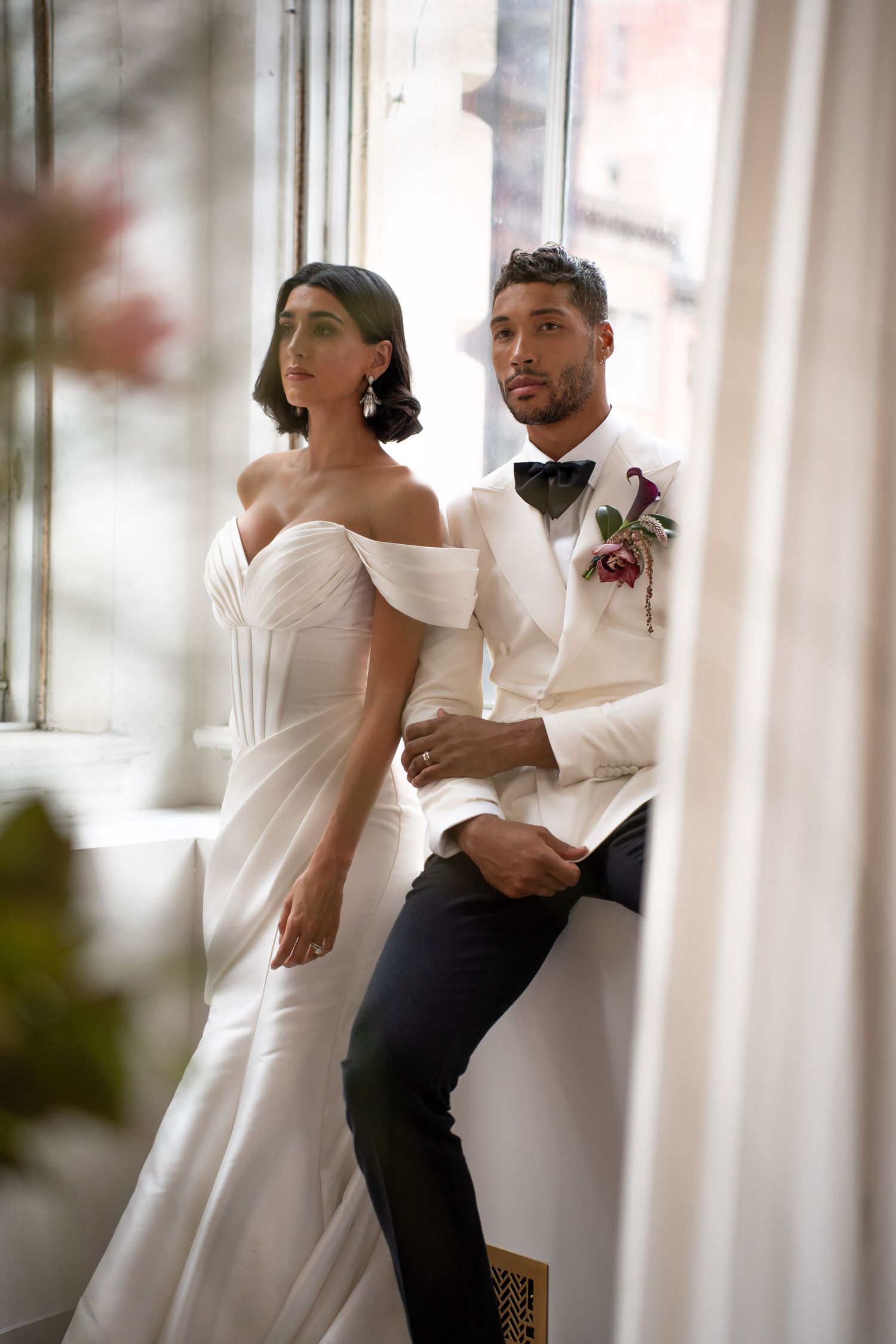 Bride in off-shoulder white gown and groom in white tuxedo with floral boutonniere posing by window.