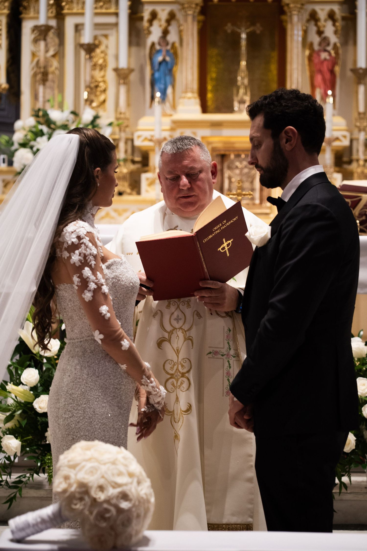 Priest reads from red Catholic wedding book as bride and groom hold hands at altar.