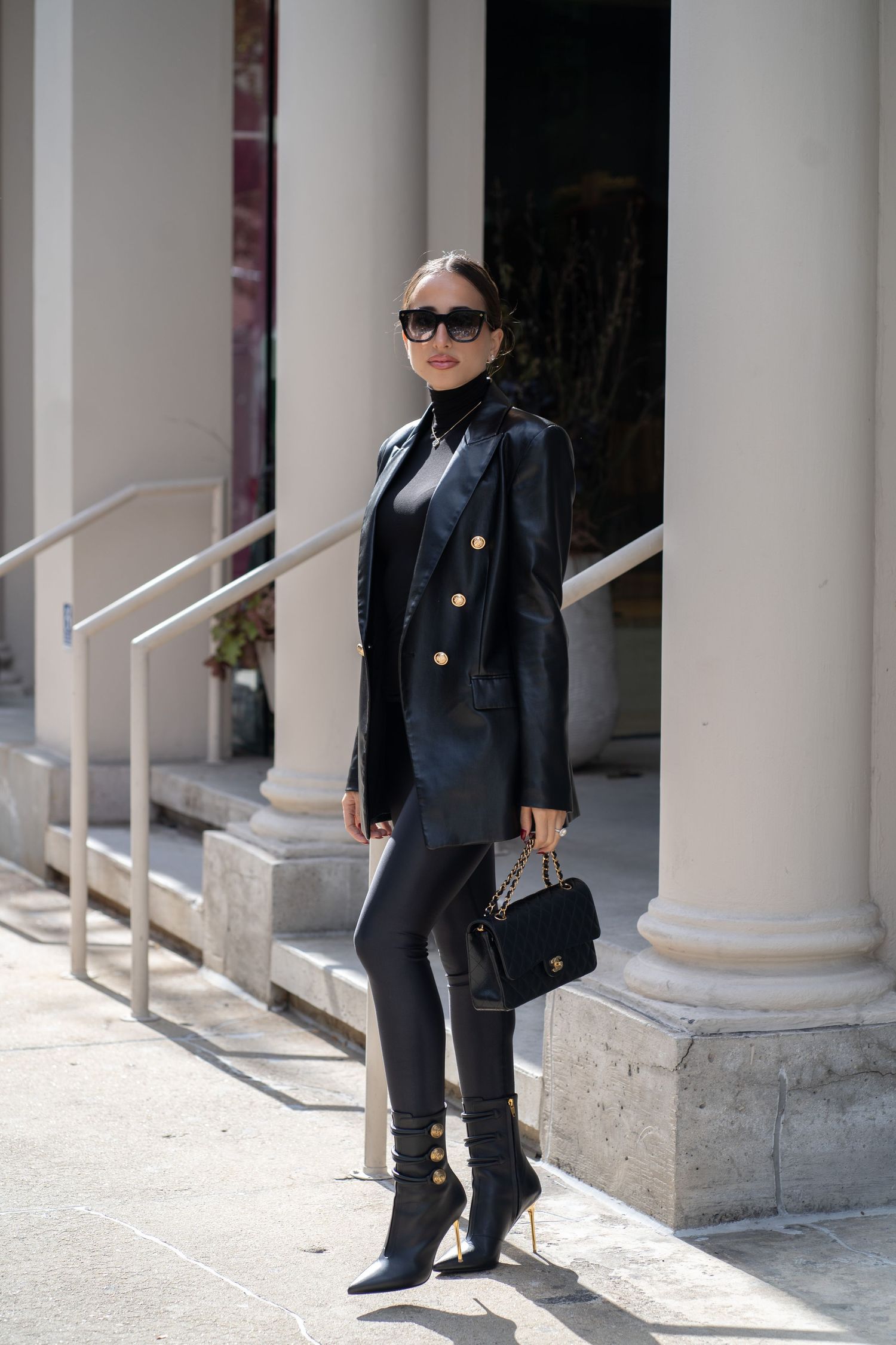 Stylish woman in black leather double-breasted blazer and leggings posing near white columns with a Chanel bag.