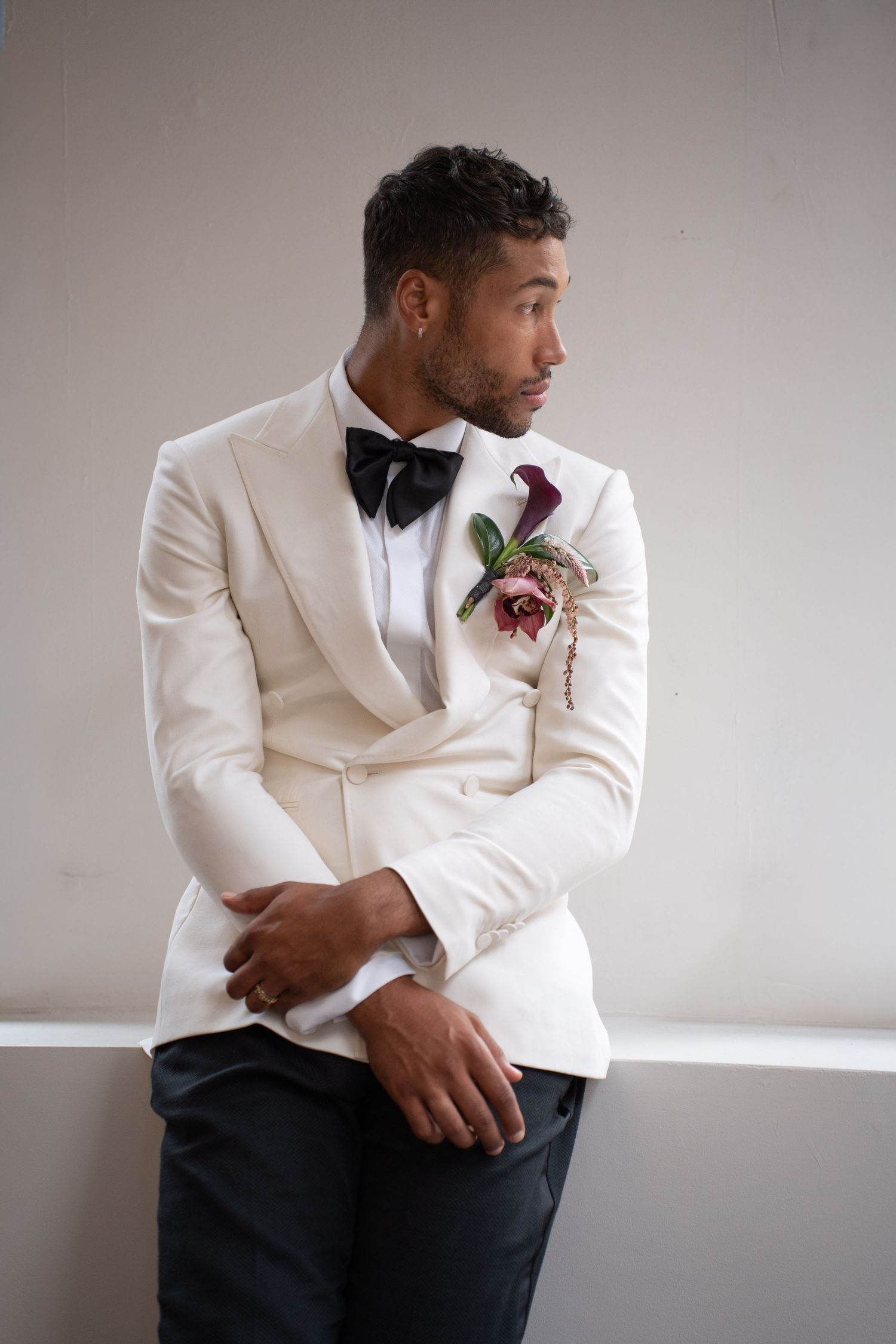 Groom in ivory double-breasted tuxedo with floral boutonniere poses against neutral background.