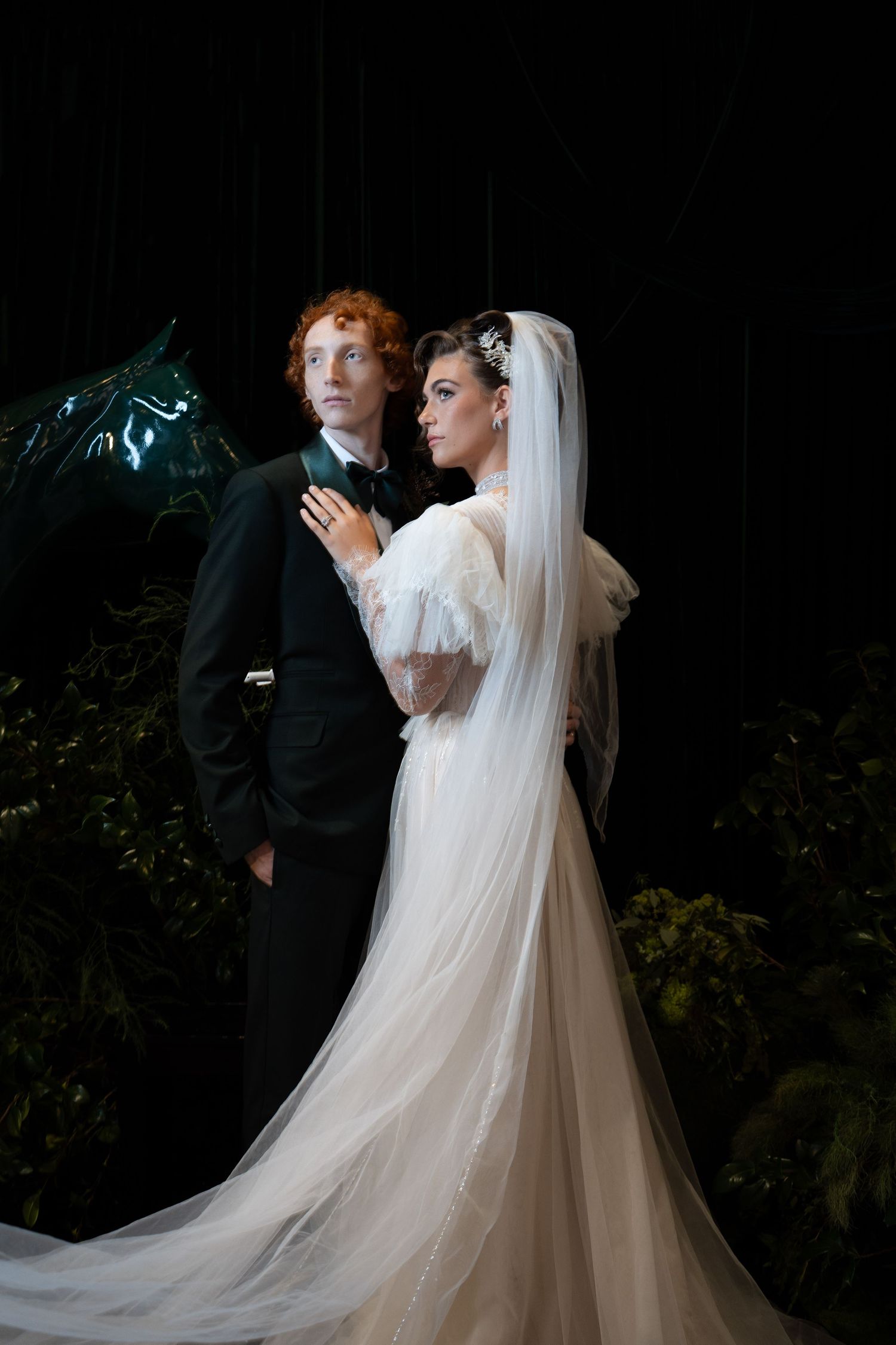 Elegant bride in tulle gown with long veil stands beside red-haired groom in black tuxedo amid dark greenery.