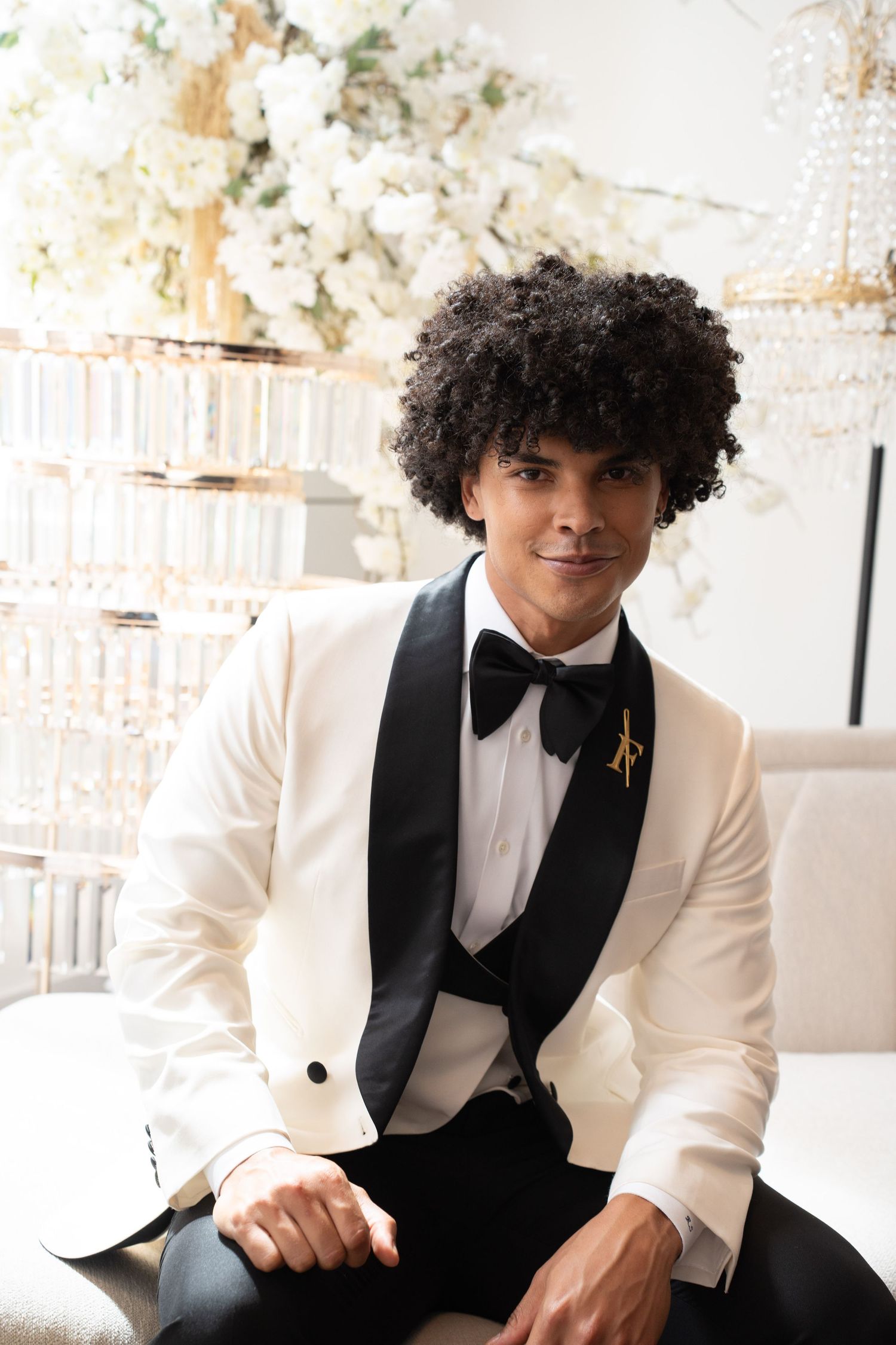 Stylish groom in white tuxedo with black bow tie and gold pin poses at elegant floral wedding venue.