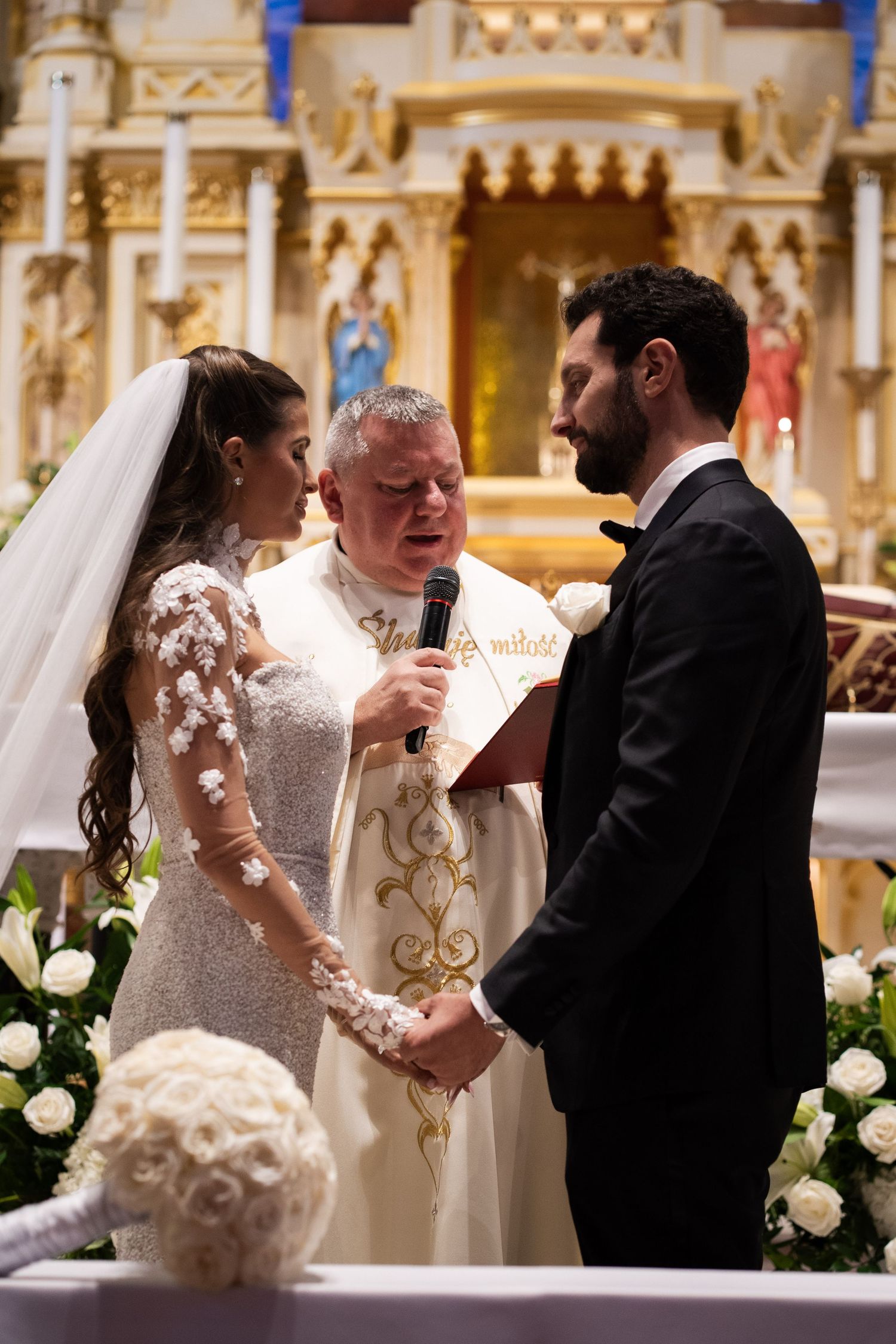 Priest reads wedding vows as bride and groom hold hands at elegant Catholic church altar ceremony.