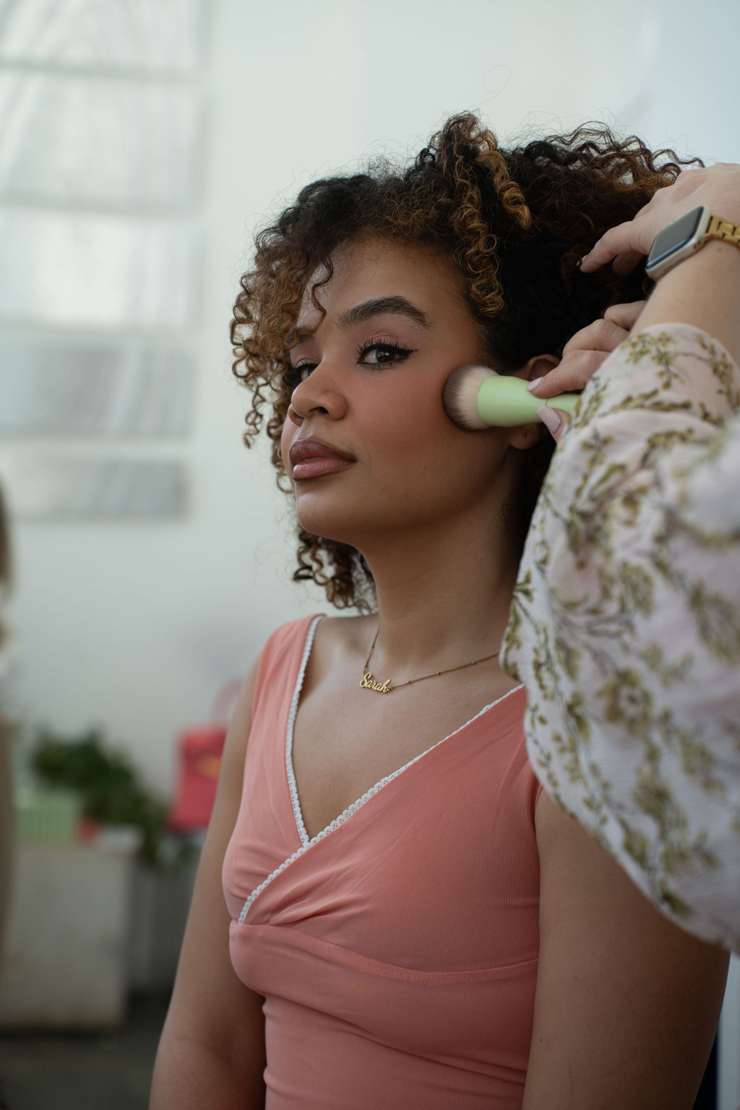 A makeup artist applies blush to a young woman with curly hair wearing a pink dress.