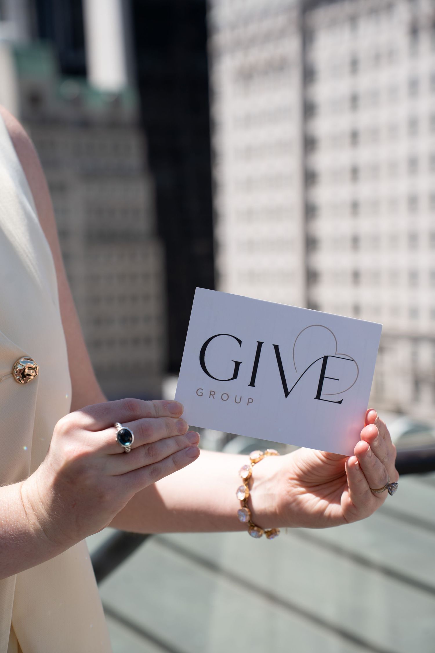 Woman holding GIVE Group branded card with pearl bracelet and ring on NYC rooftop terrace.