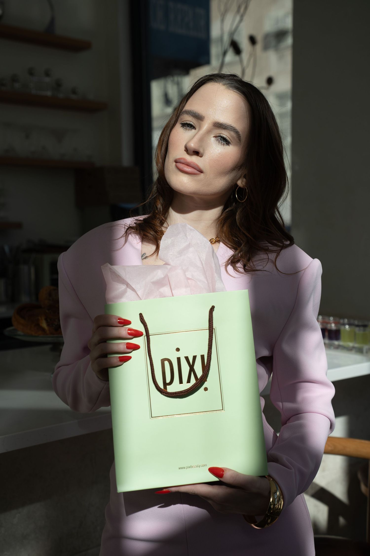 Brunette woman in pink outfit holding a mint green Pixi beauty shopping bag indoors.