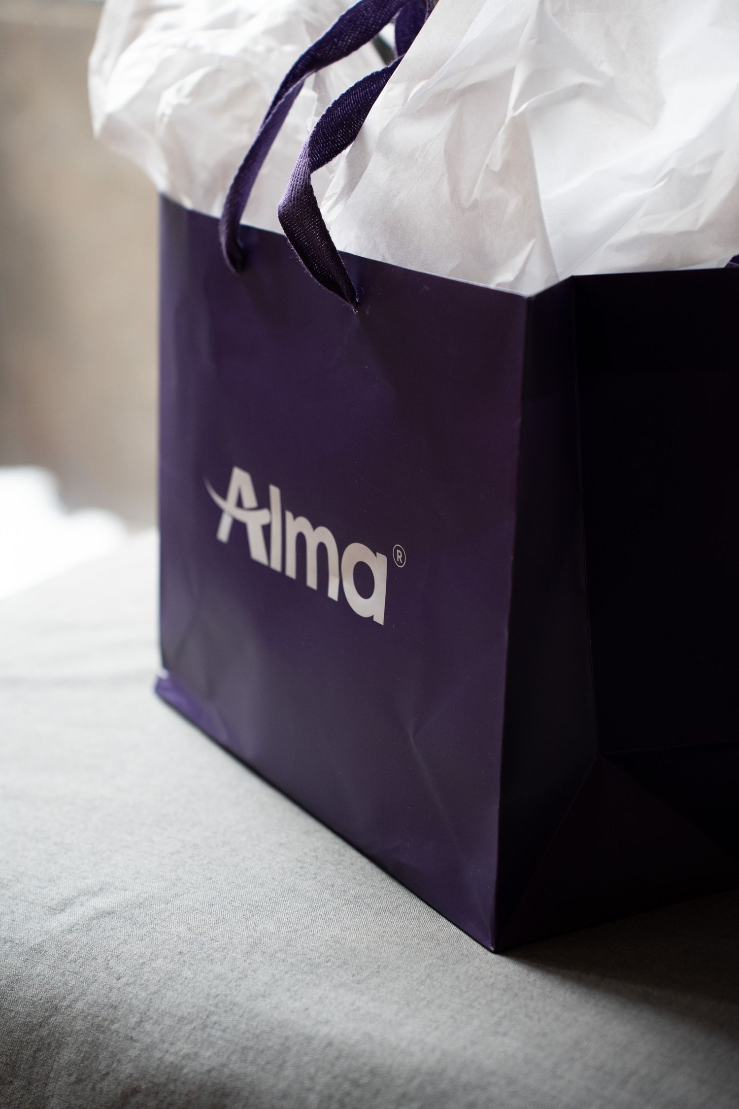 Close-up of a purple Alma laser aesthetics branded shopping bag with white tissue paper.