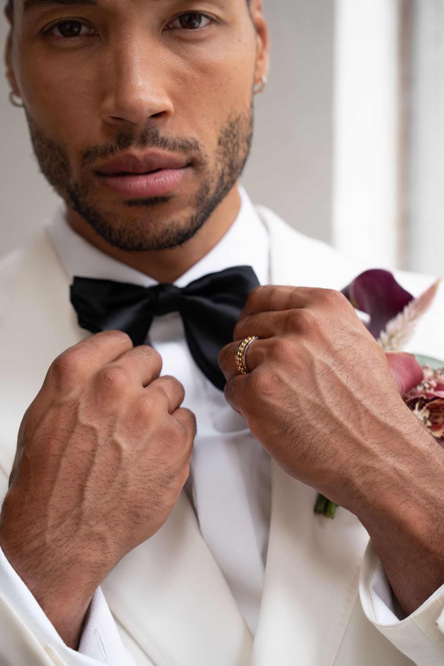 Close-up of groom in white tuxedo adjusting black bow tie, wearing gold ring with burgundy boutonniere.