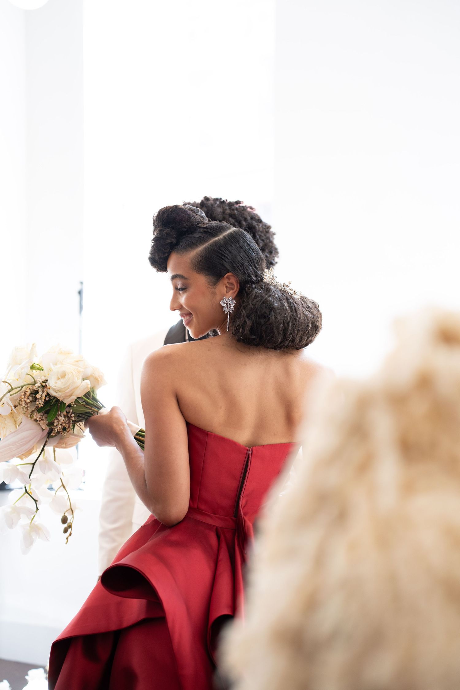 Elegant woman in red strapless dress with updo and chandelier earrings admires white bouquet, photographed from behind.