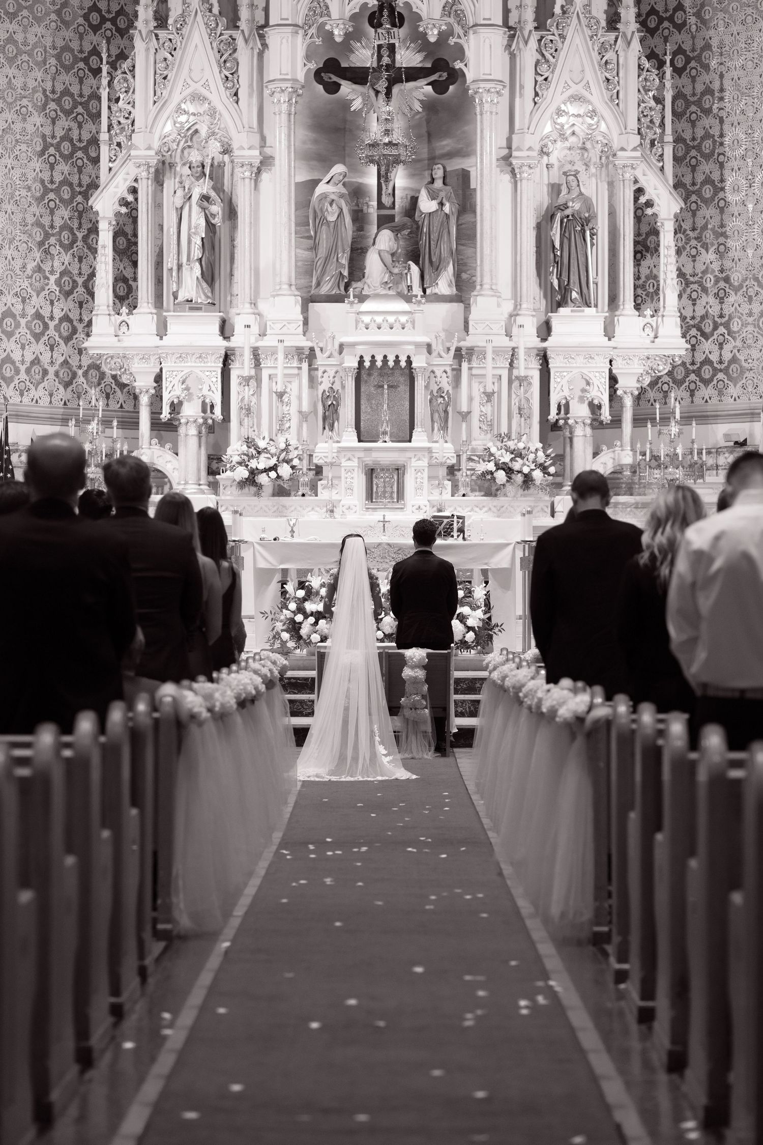 Bride and groom kneel at the altar of a stunning Catholic church adorned with statues, candles, and floral arrangements.