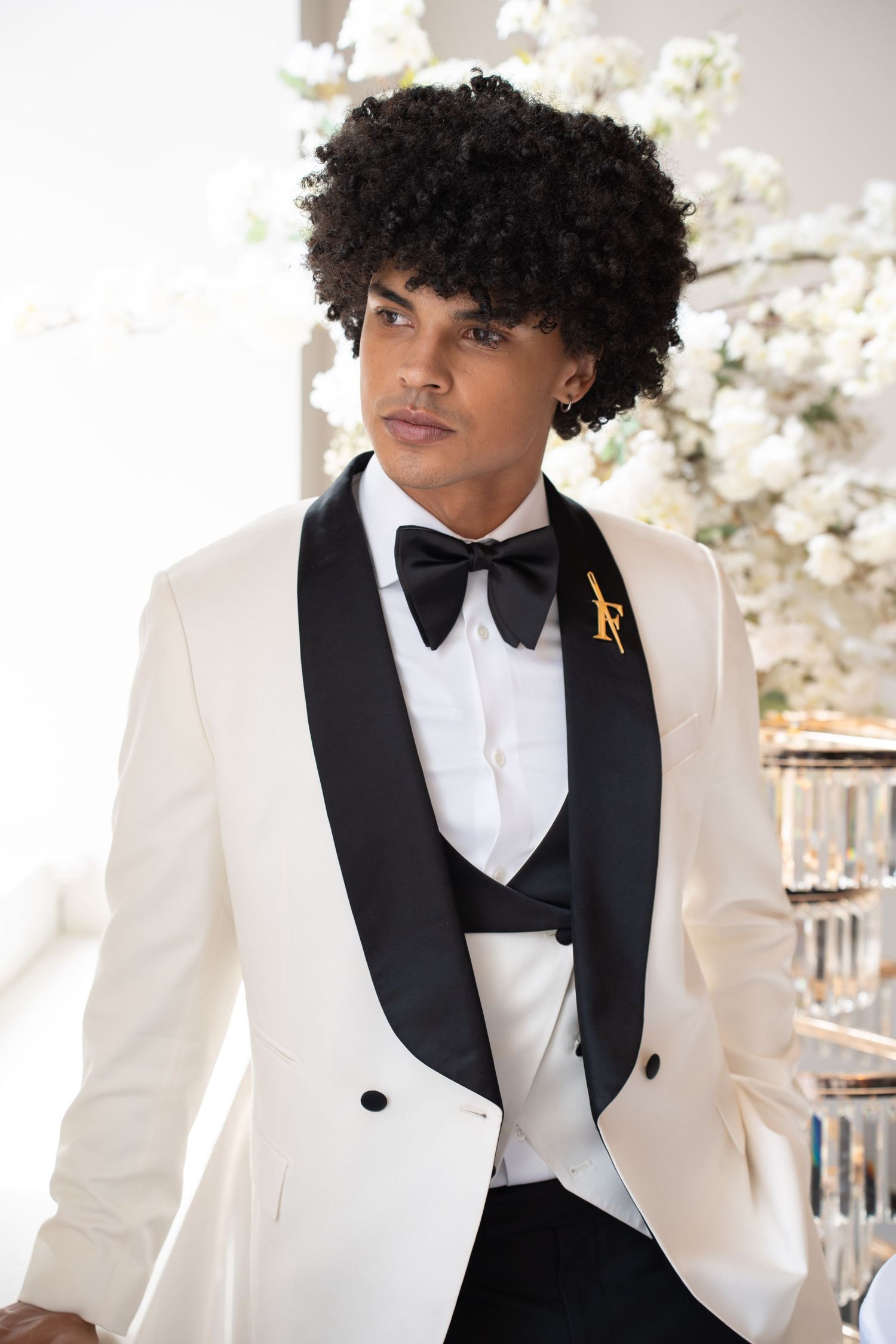 Handsome groom in white shawl lapel tuxedo with black bow tie and gold monogram brooch looking away.