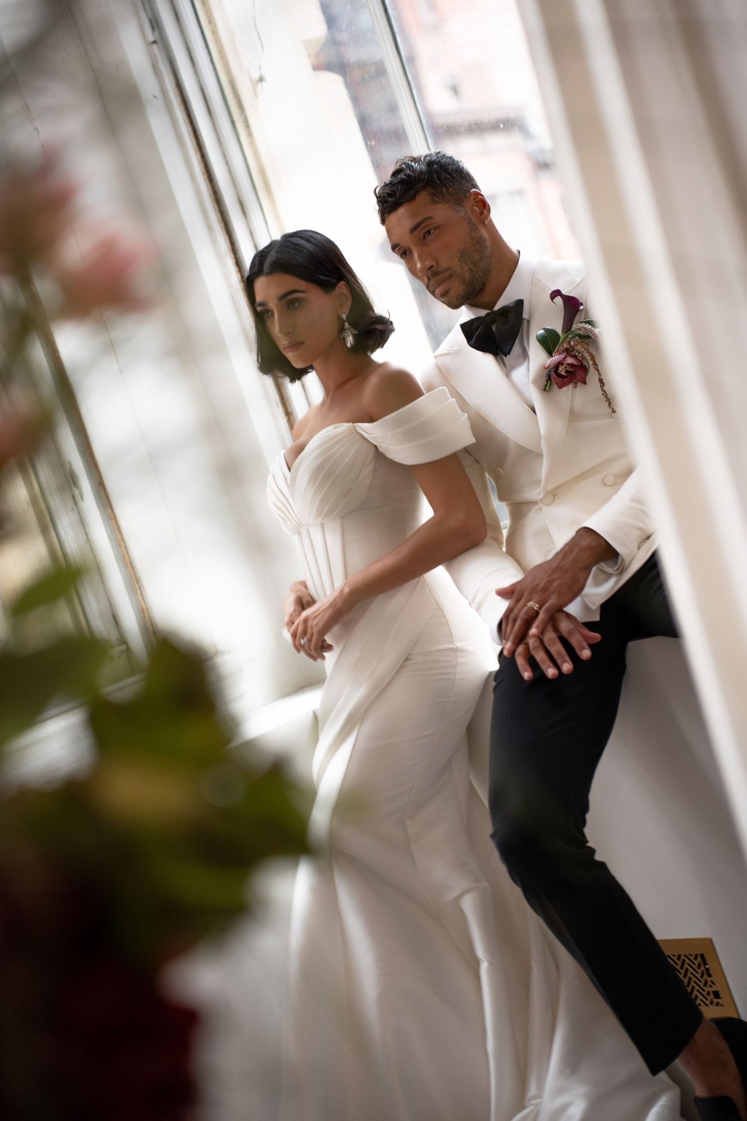 Bride and groom in coordinating white wedding attire pose together by sunlit window.