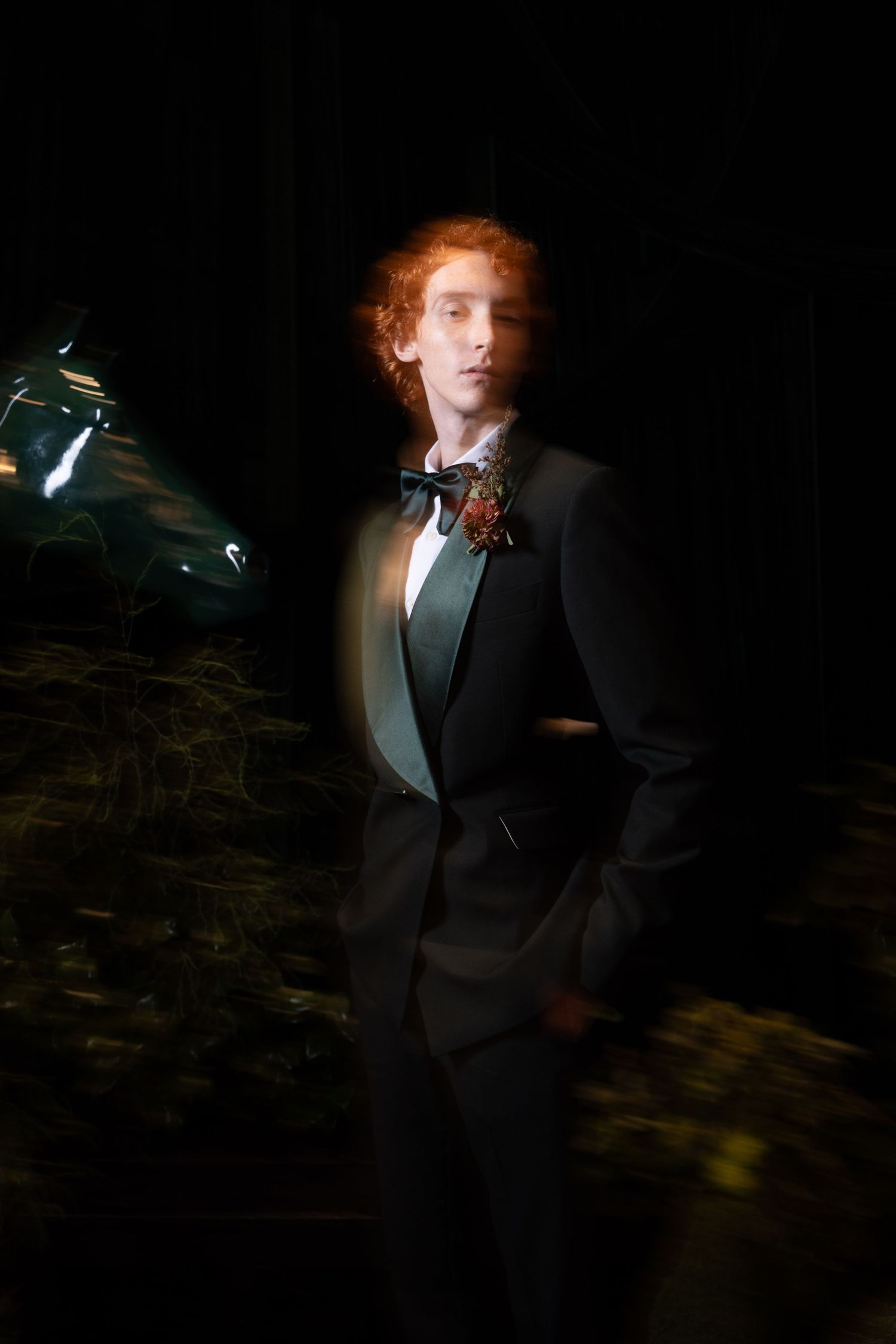 Red-haired groom in black tuxedo with floral boutonniere poses solo in artistic motion-blur photograph.