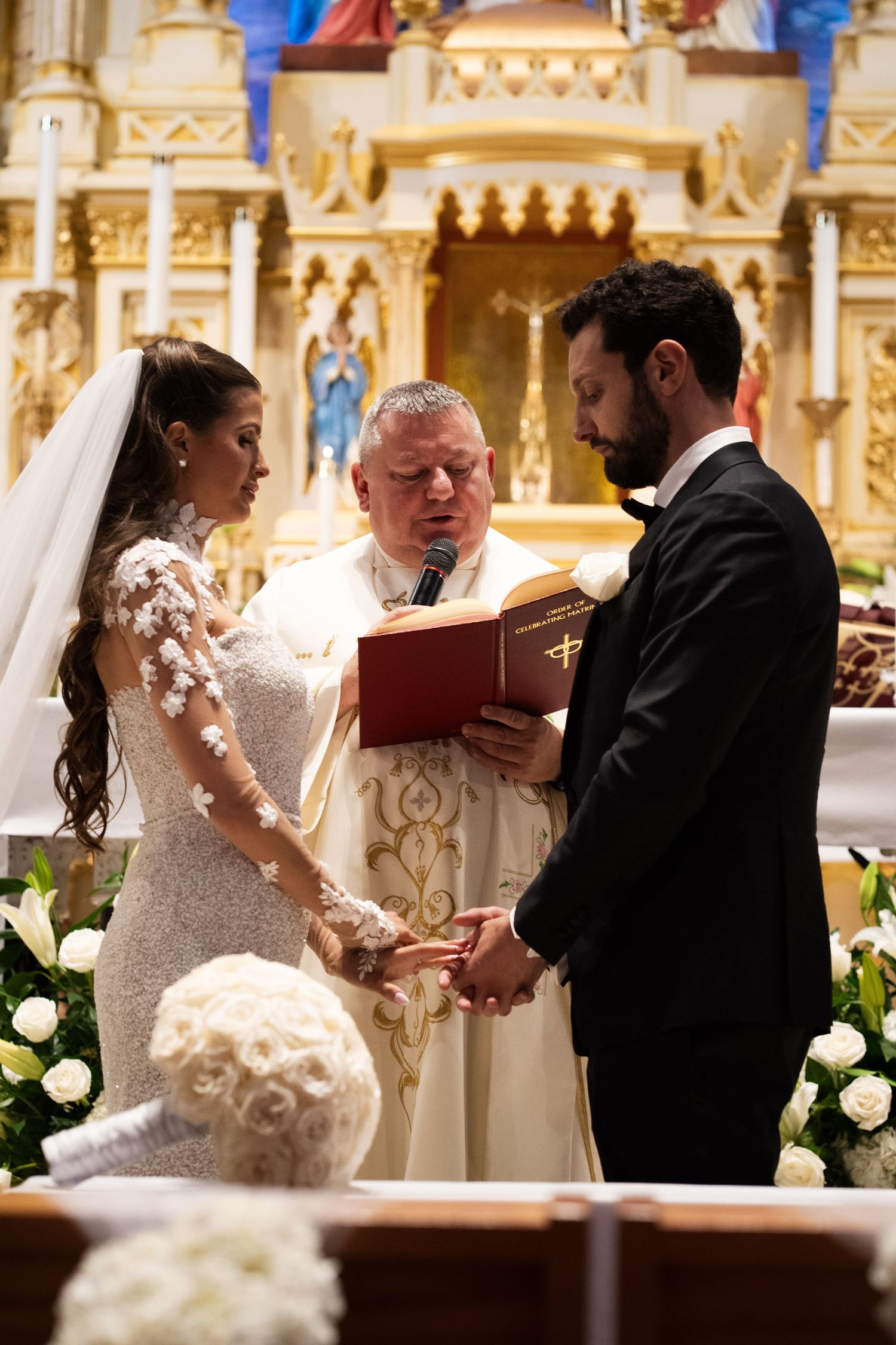 Priest reads from red Bible as bride and groom hold hands during Catholic wedding ceremony.