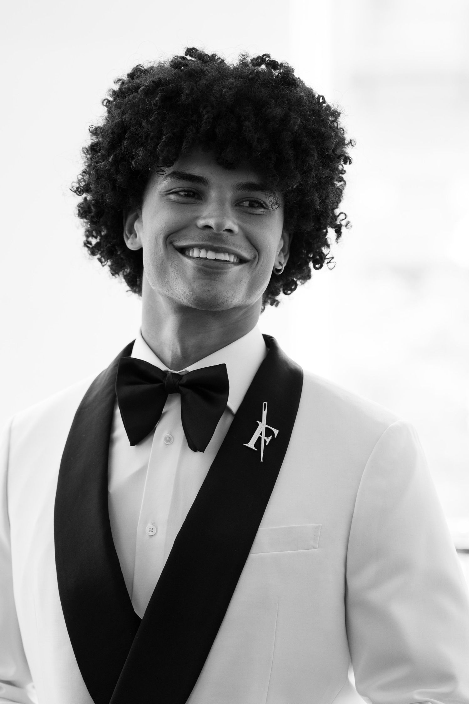 Black and white portrait of smiling groom in white tuxedo with black bow tie and decorative lapel pin.