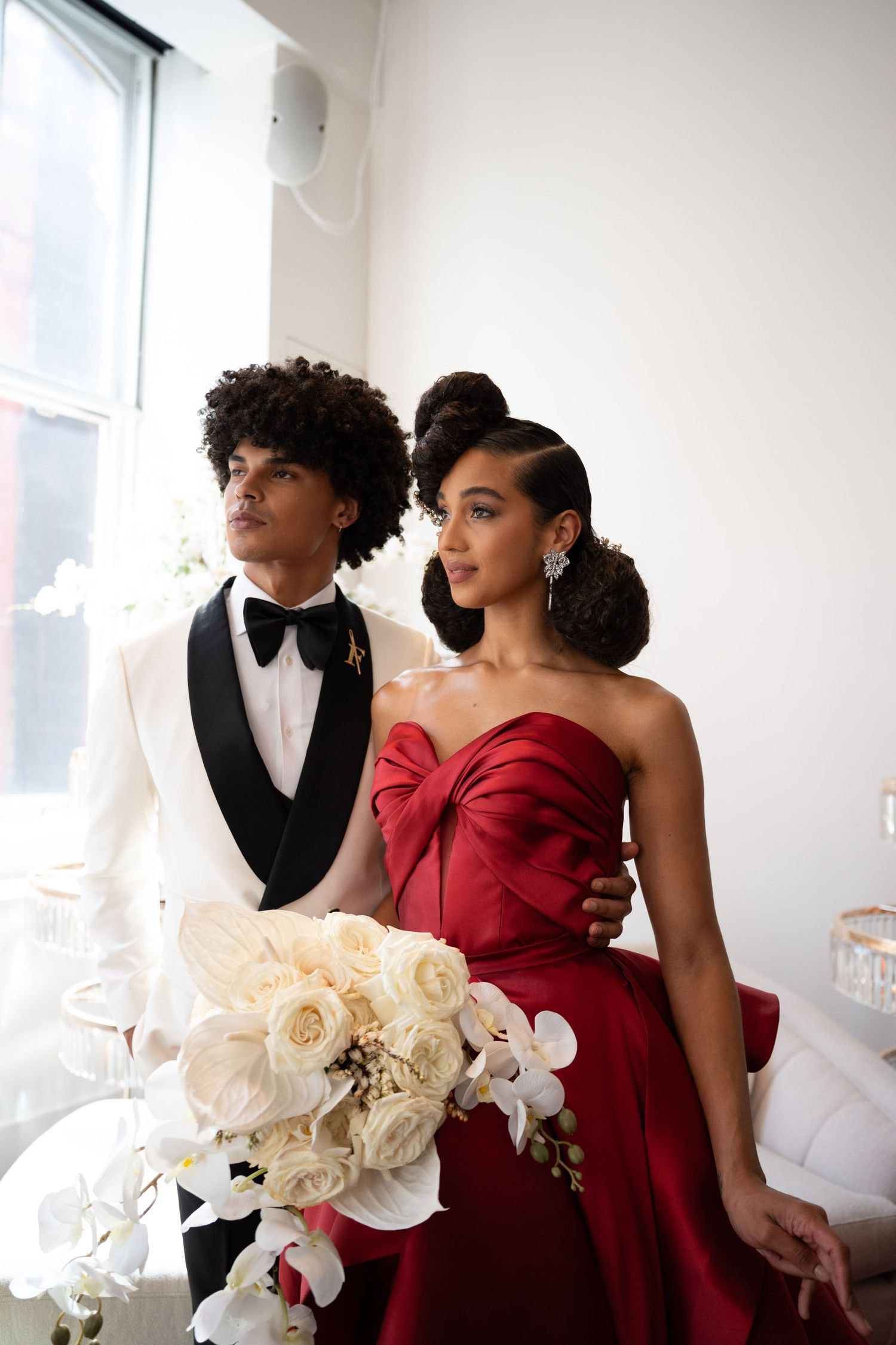 Bride in red strapless ballgown and groom in white tuxedo posing with white floral bouquet.
