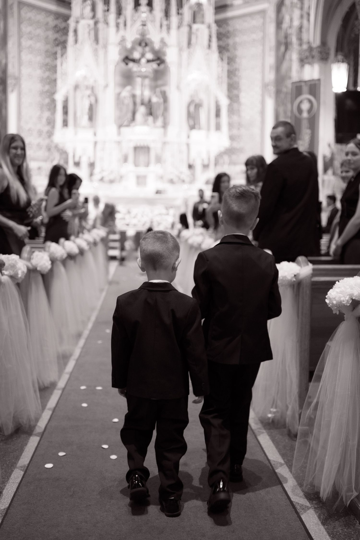 Two young ring bearers in black suits walk hand-in-hand down the aisle of an ornate Catholic church during a wedding.
