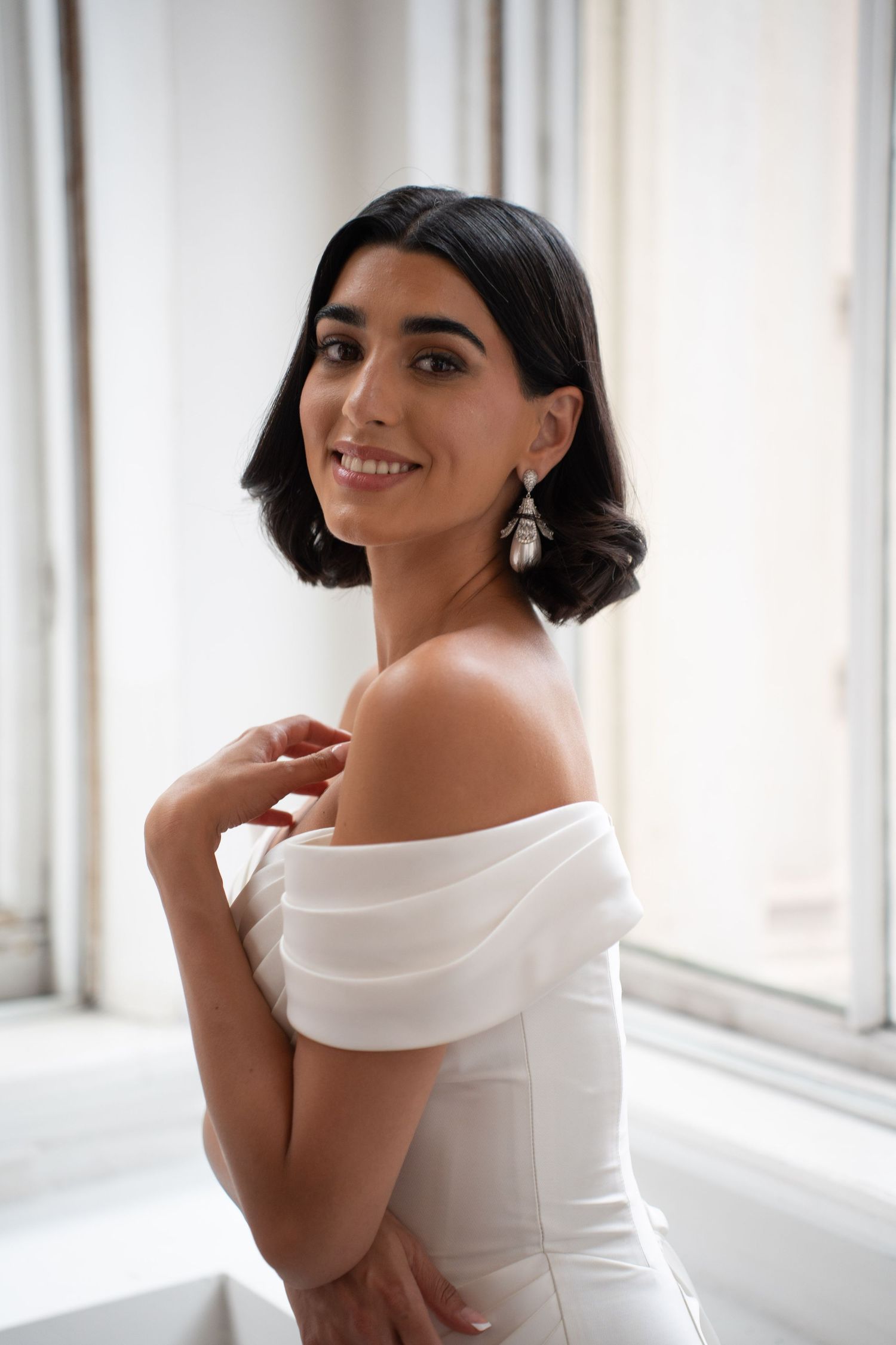 Smiling bride in off-shoulder white corset gown touches her shoulder while wearing pearl drop earrings indoors.