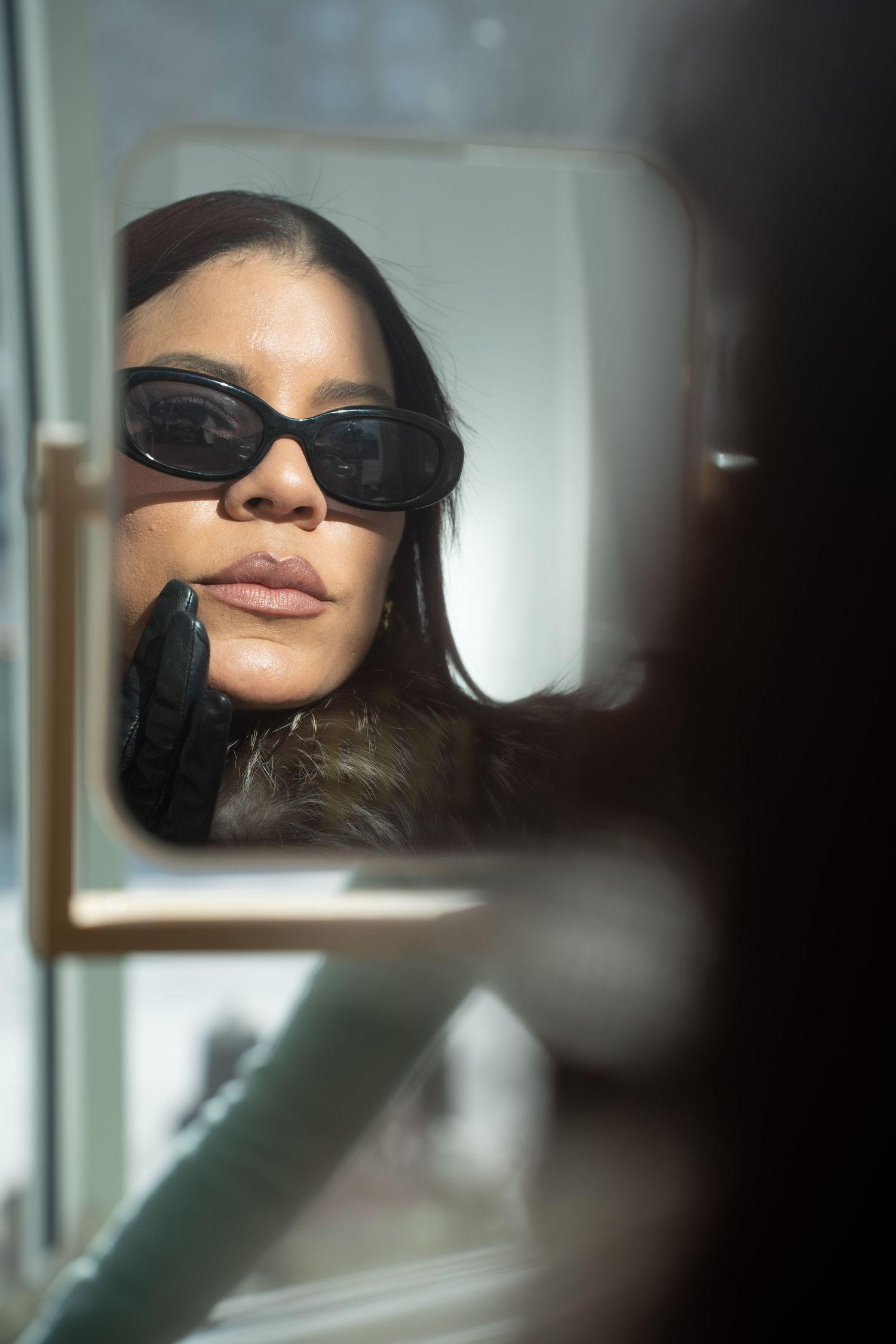 Woman with dark sunglasses and black glove examining her reflection in a small handheld mirror.