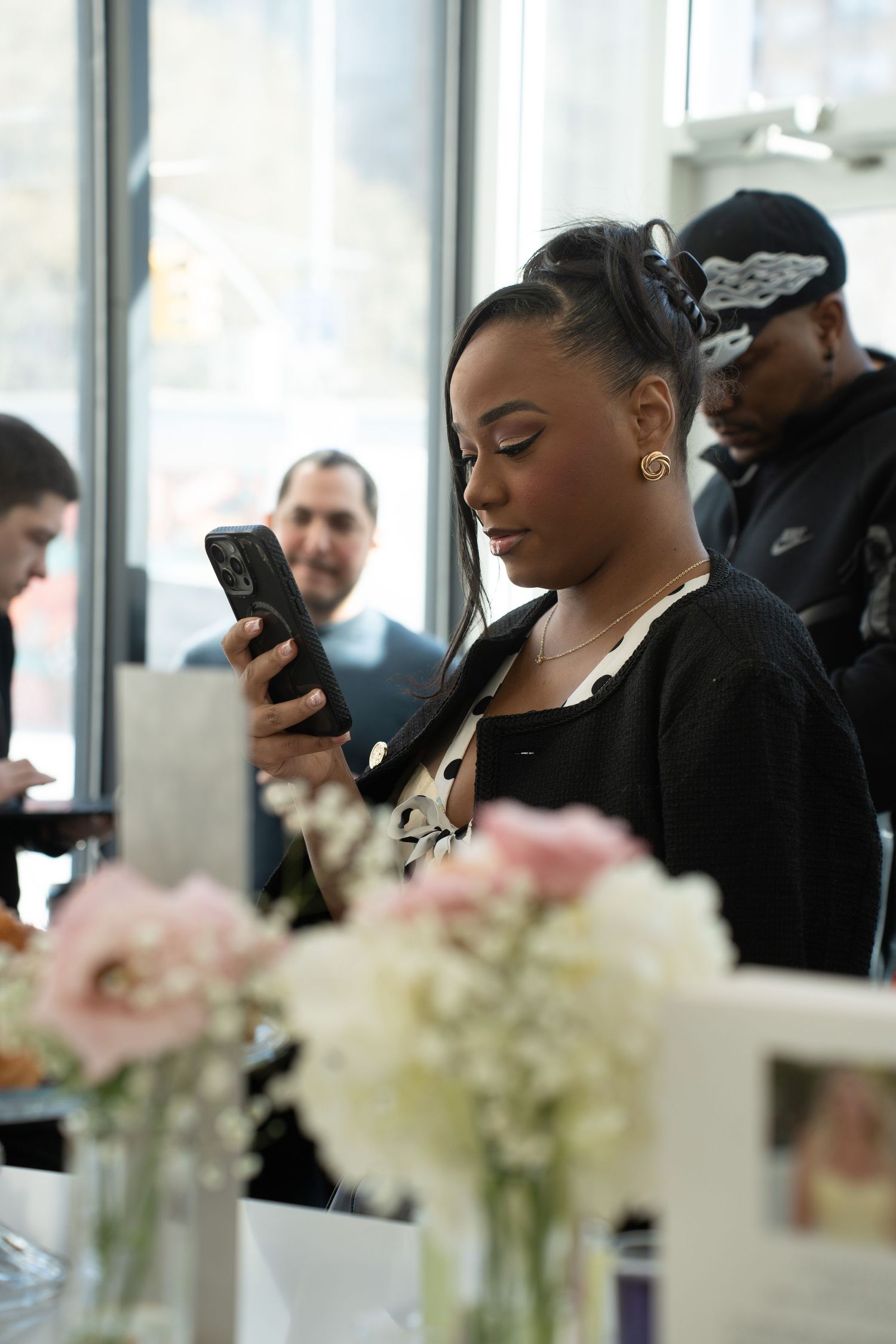 Young woman in black outfit looking at her smartphone at an indoor event with floral decorations.
