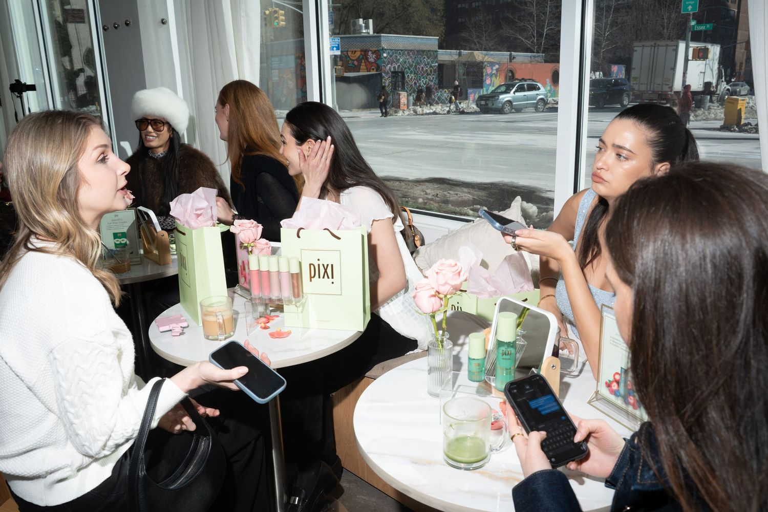 Women sitting at a cafe table with Pixi beauty bags and green drinks, using smartphones.